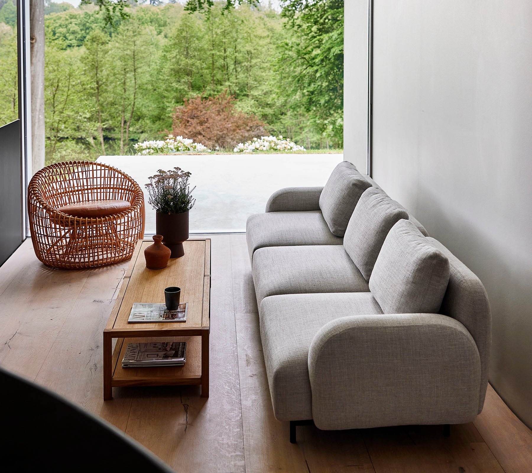 Moderne bank in lichte stof, gecombineerd met een houten salontafel en een rotanstoel, omringd door groen. Natuurlijk zonlicht verbetert de serene ambiance.