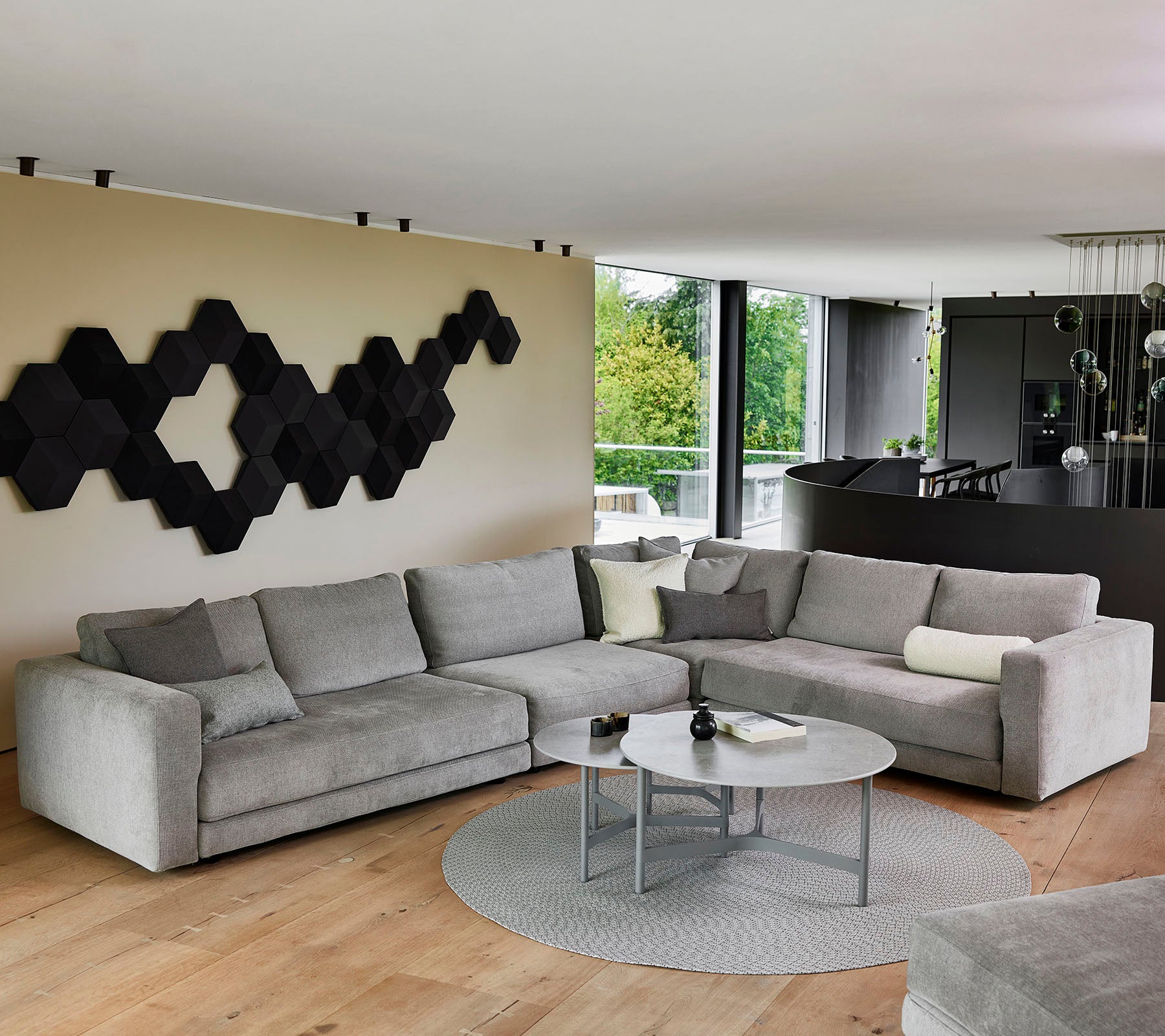Een moderne grijze hoekbank met decoratieve kussens, een ronde salontafel en een gestructureerd zwart wandkunstwerk.