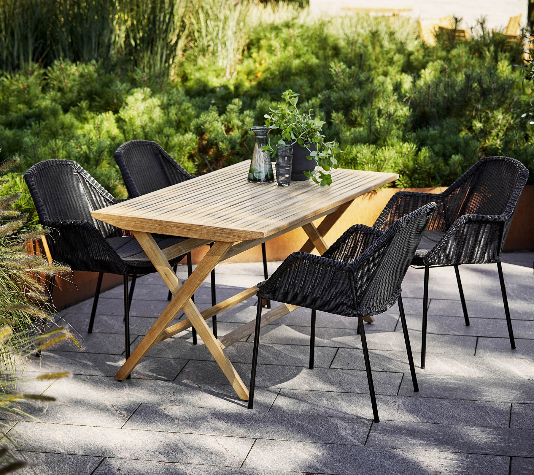 Een houten eettafel omringd door vier zwarte gevlochten stoelen, met groen in de achtergrond en decoratieve items op de tafel.