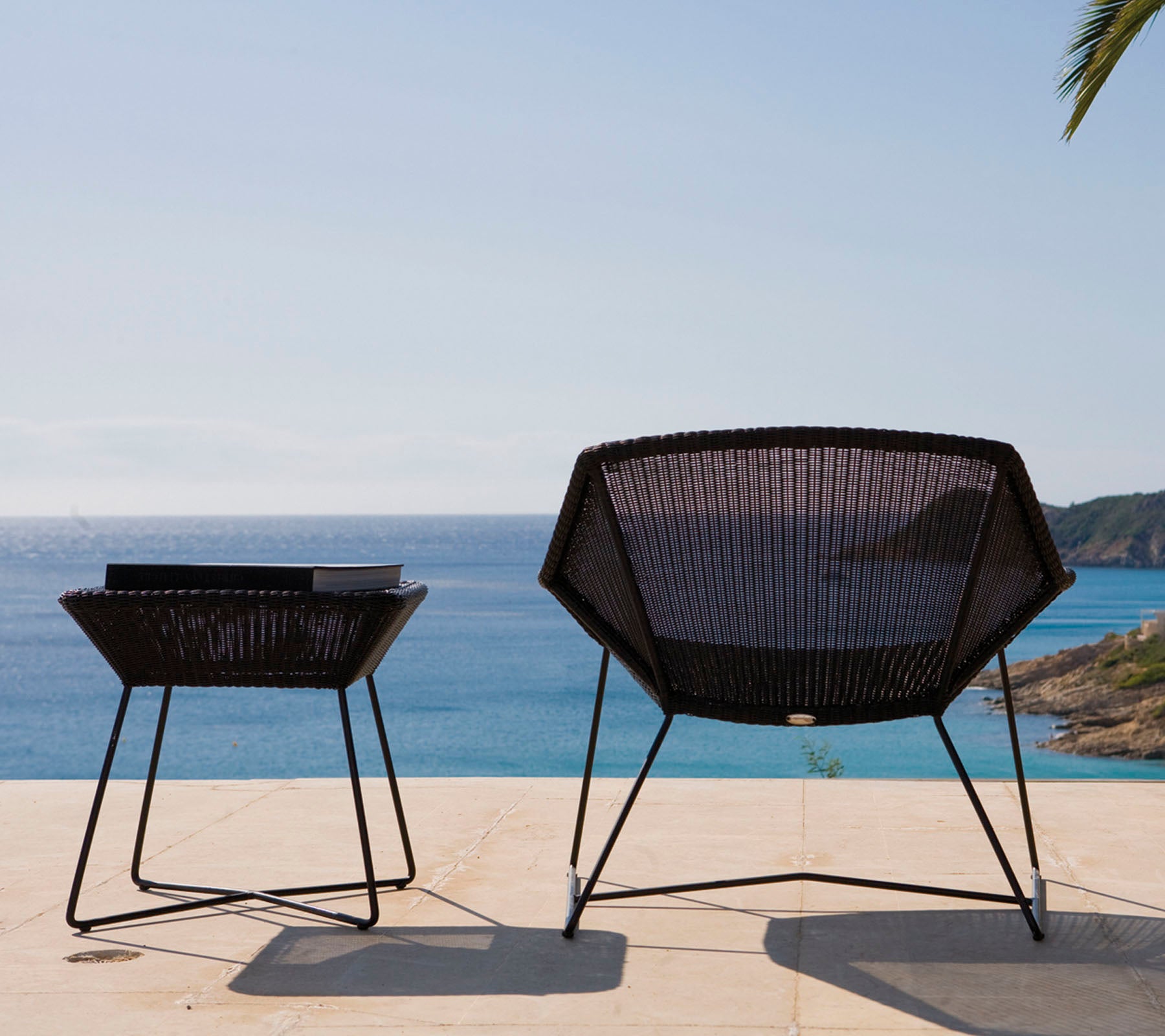 Twee moderne, zwarte tuinstoelen met een geometrisch design staan naast een tafel en kijken uit over een kalme zee onder een blauwe lucht.