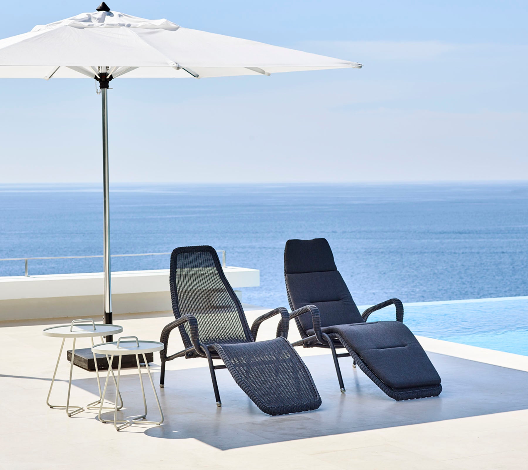 Twee stijlvolle loungestoelen en een klein tafeltje staan onder een grote parasol, met een sereen blauw zee op de achtergrond.