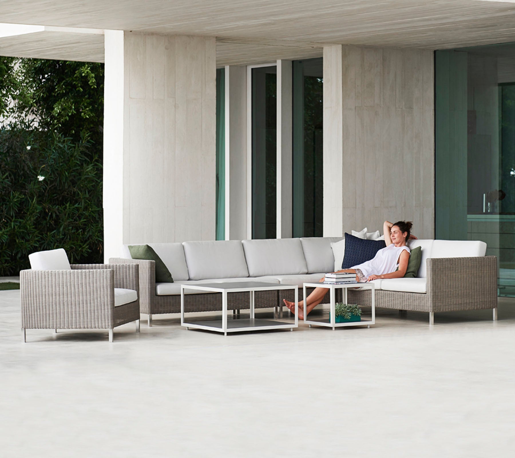 Een moderne zithoek met een grijze hoekbank, een glazen tafel en een rotan stoel, met een persoon die comfortabel ontspant.