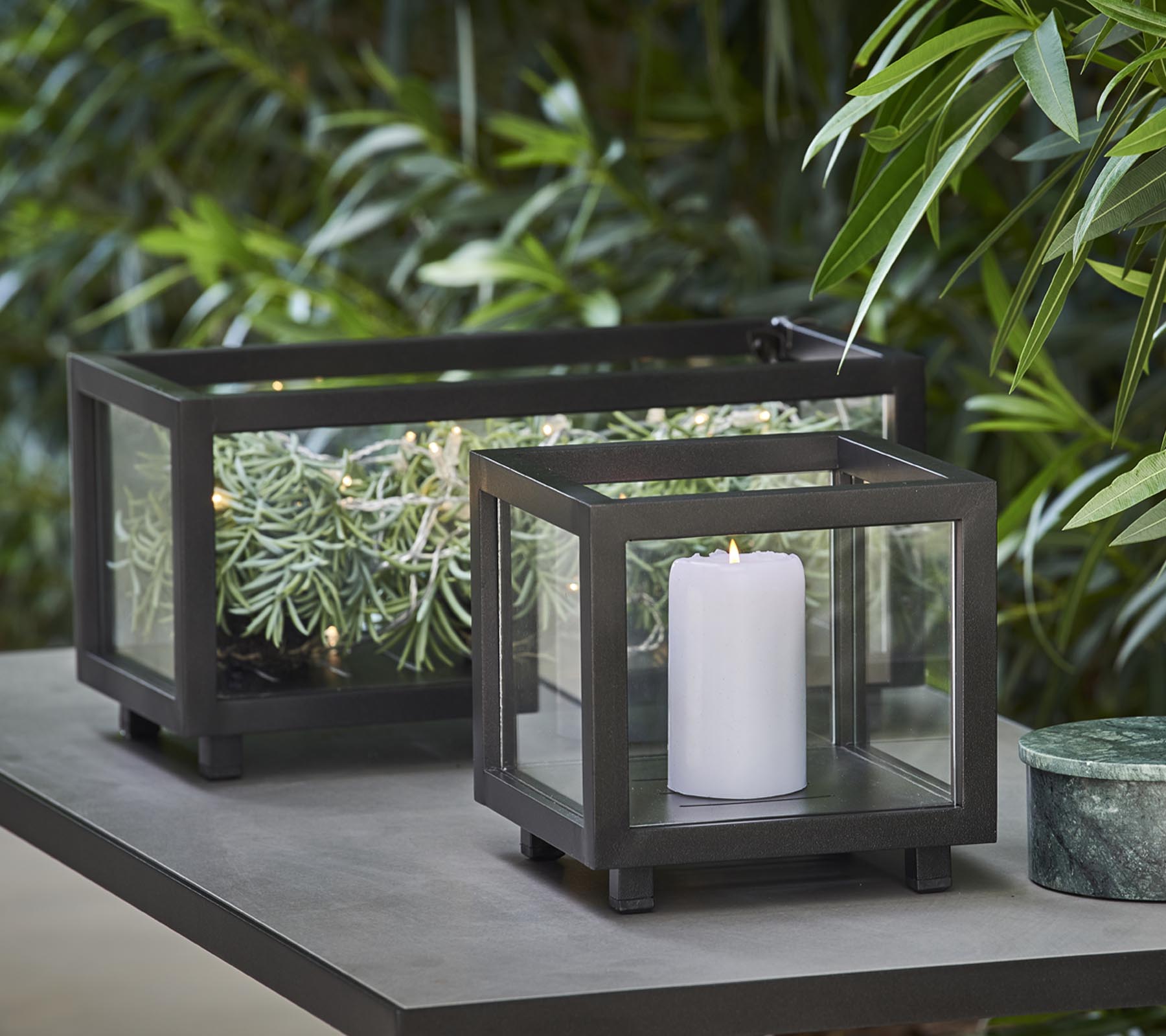 Twee stijlvolle lantaarns met glazen panelen, de ene met een witte kaars en de andere met groen, geplaatst op een tafel te midden van weelderig loof.