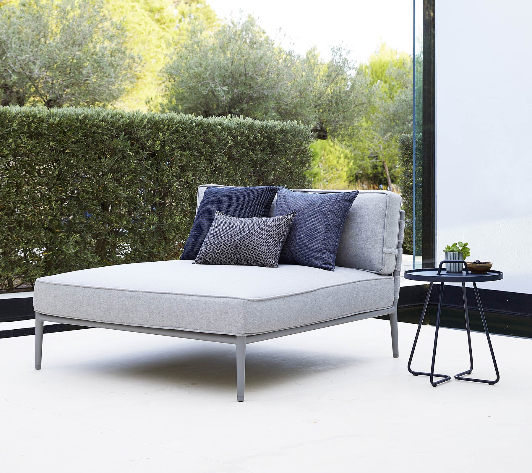 Stijlvolle grijze loungechair met marineblauwe kussens naast een klein zwart bijzettafeltje in een rustige omgeving.