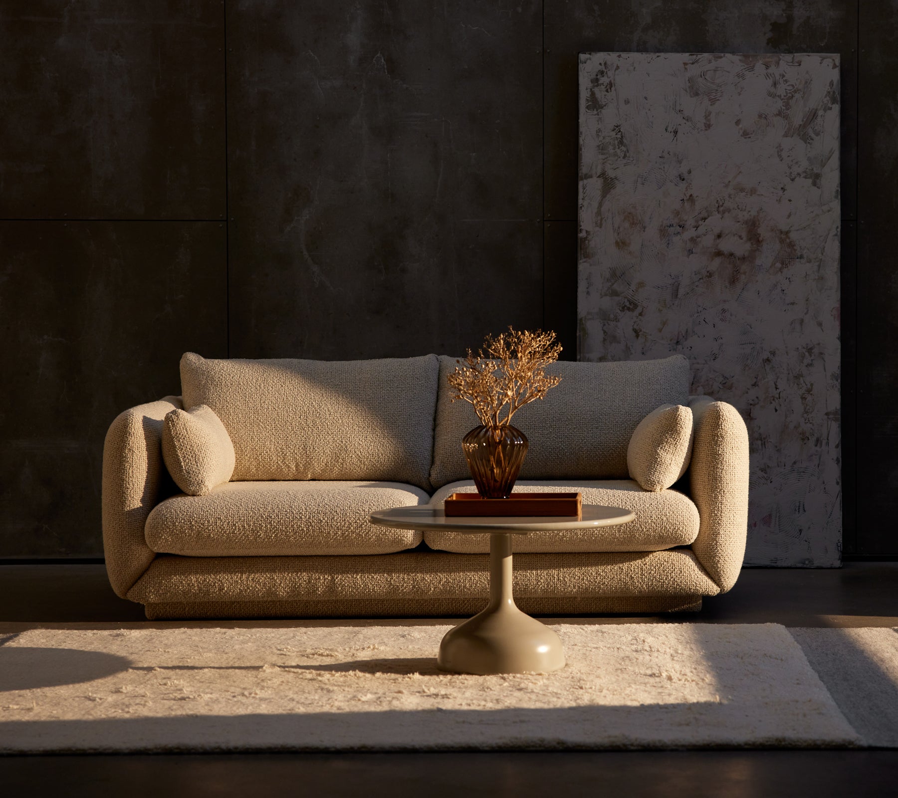 Bloom 2-zitsbank in zand flair met een Glaze salontafel en een decoratieve plant op een tapijt, geplaatst tegen een gestructureerde donkere muur.