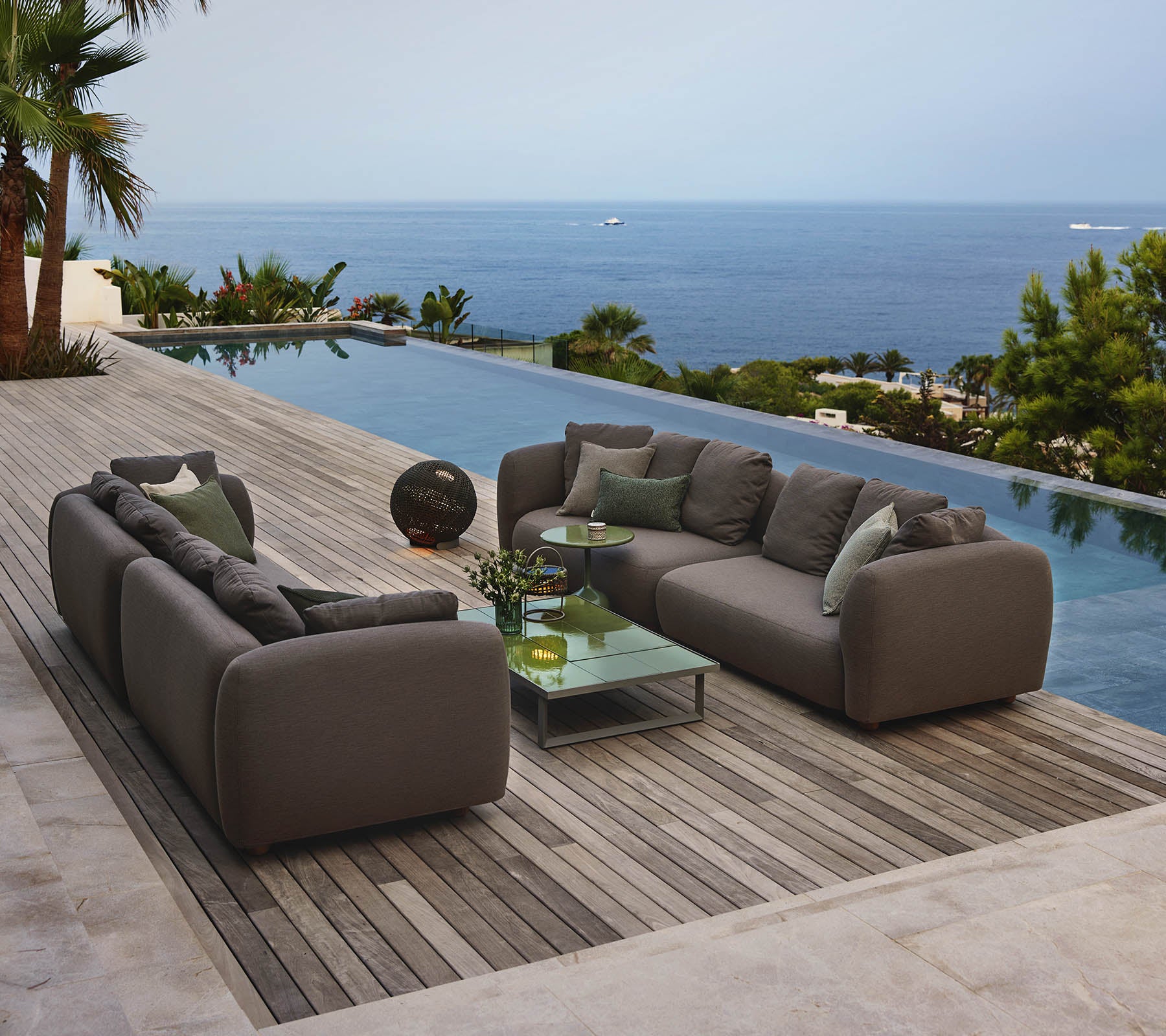 Moderne loungezitplaatsen rond een glazen salontafel, met uitzicht op een serene pool en een uitgebreid uitzicht op de oceaan, omgeven door groen.