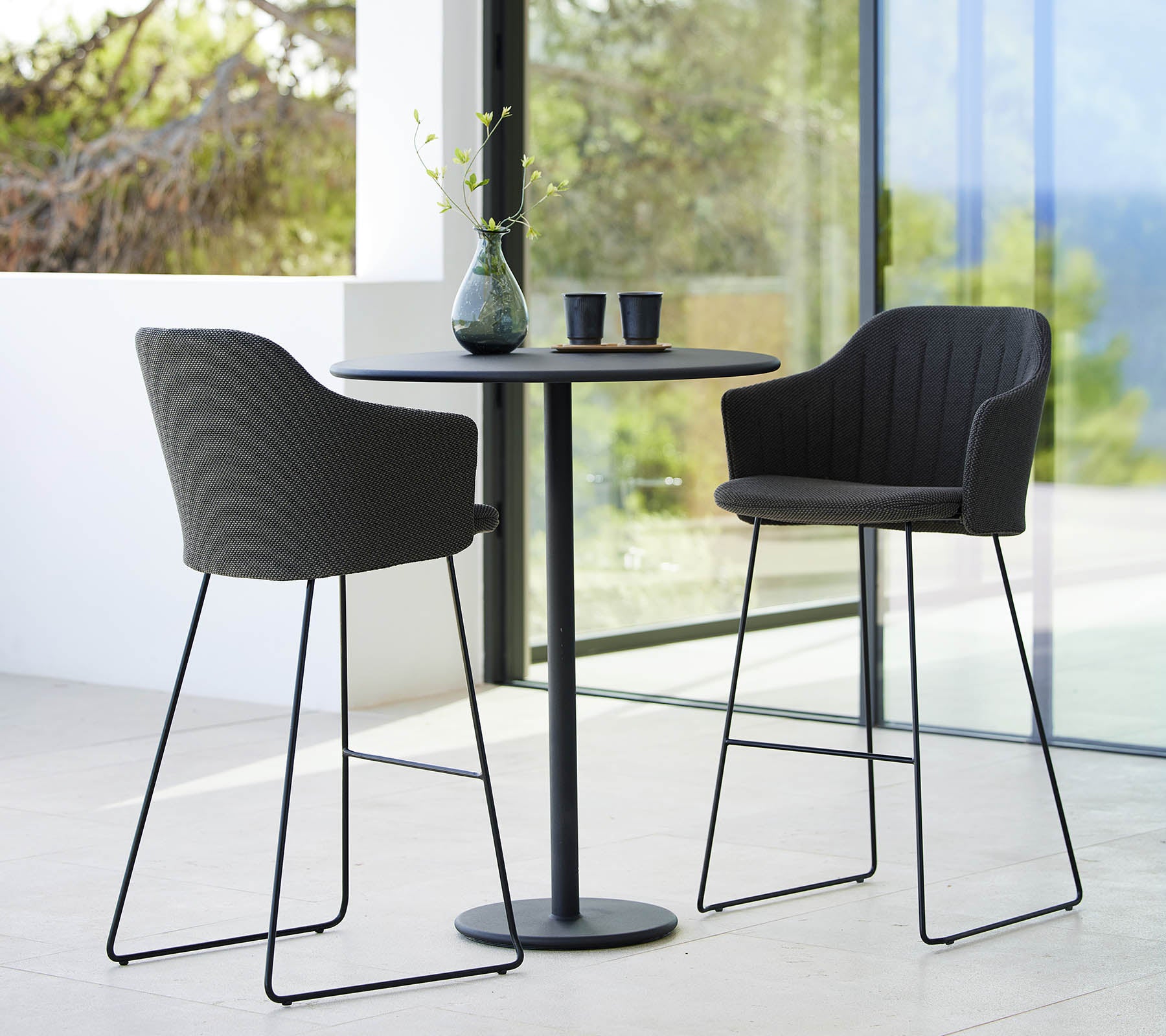 Twee stijlvolle keuze barkrukken gecombineerd met een slanke ronde bartafel, accentueert door decoratieve voorwerpen en een natuurlijke achtergrond.