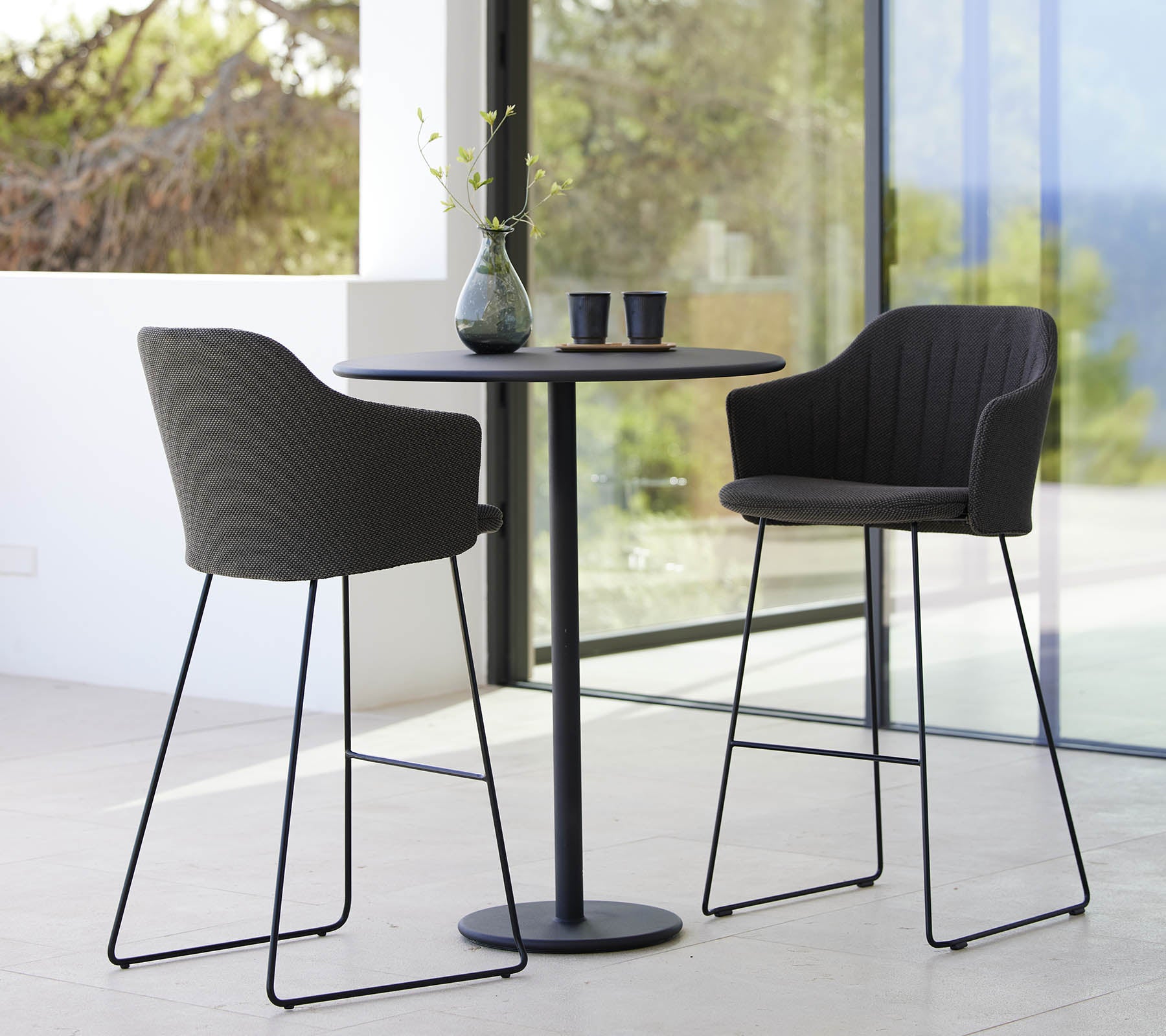 Twee stijlvolle stoelen gecombineerd met een sleek bartafel, aangevuld met een vaas en kopjes, waardoor een moderne en uitnodigende zithoek ontstaat.