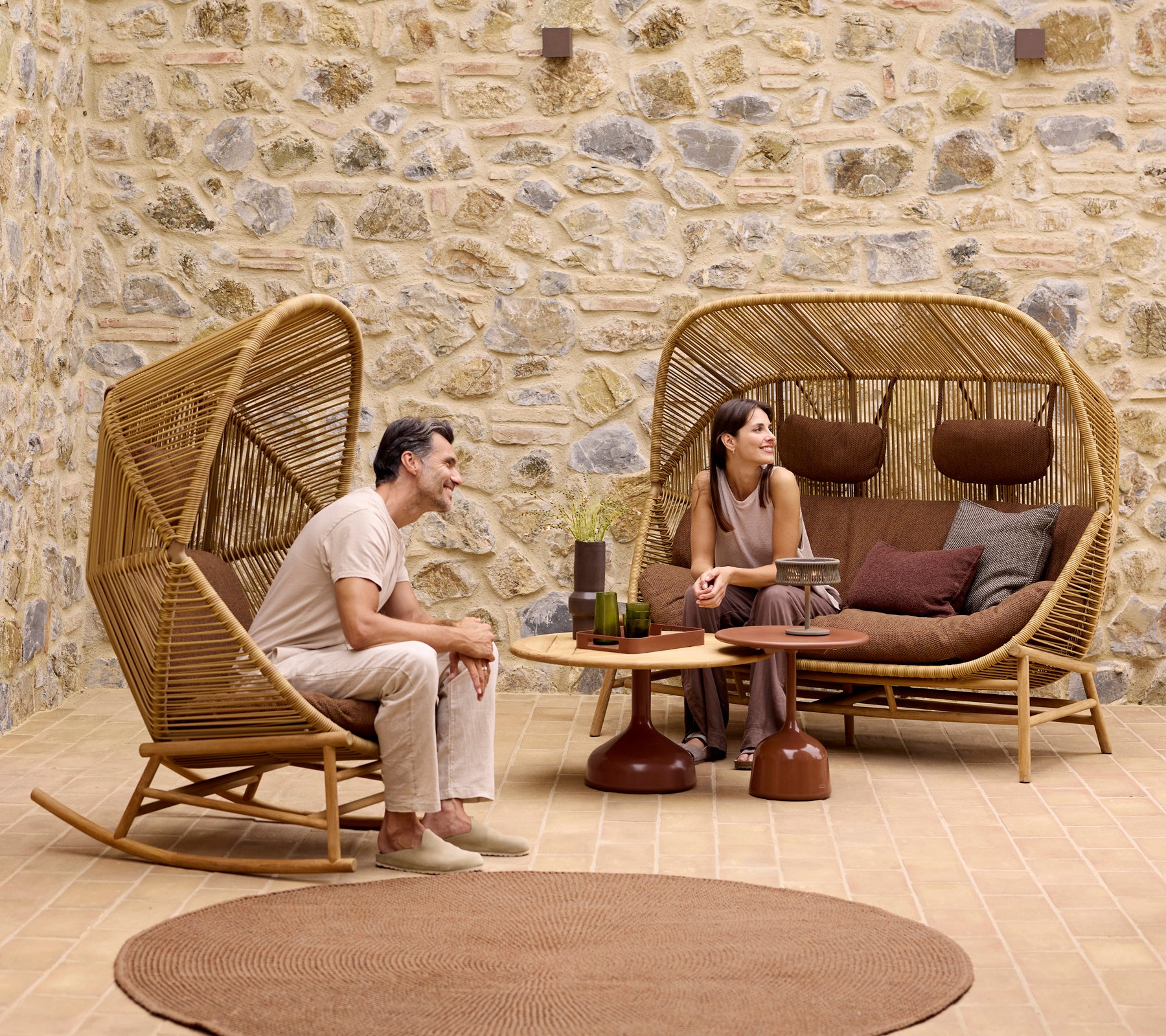 Twee mensen ontspannen in stijlvolle Hive schommelstoelen en een Hive-sofa, omringd door Glaze salontafels en een zacht gebreid tapijt.