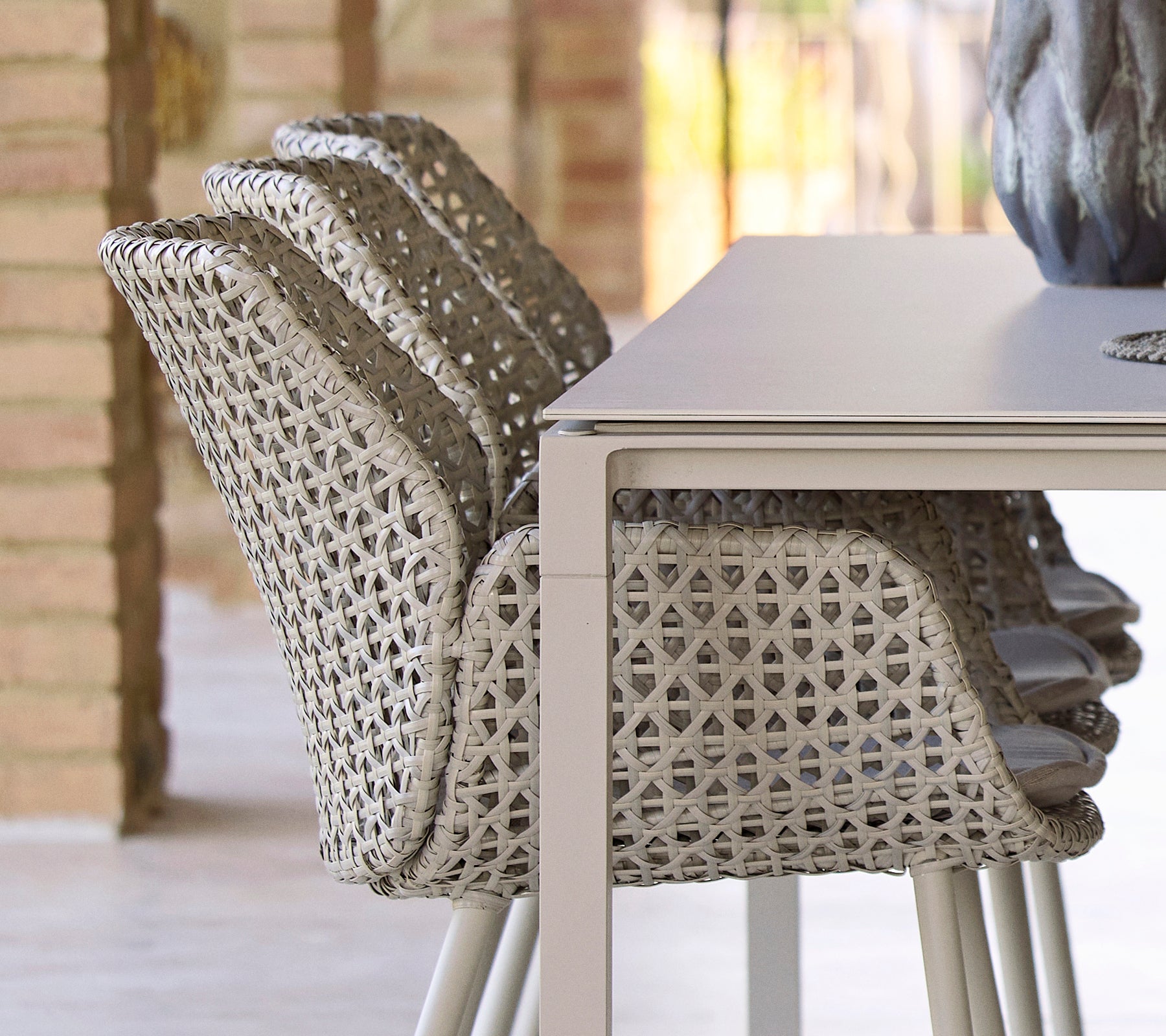 Gestapelde stoelen met een uniek geweven ontwerp naast een glad, minimalistisch eettafel, waardoor een moderne en uitnodigende eetopstelling ontstaat.