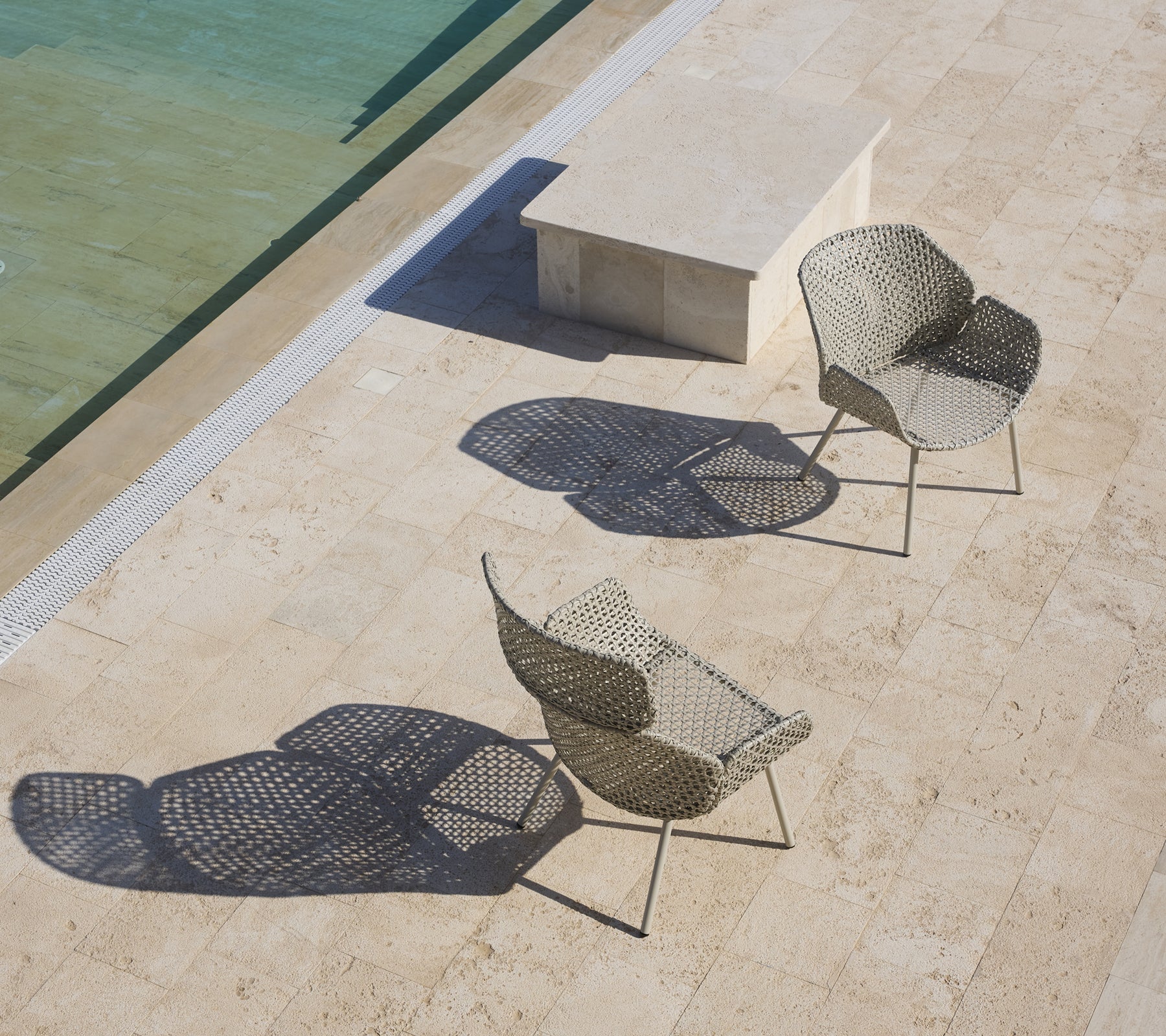 Twee stijlvolle loungestoelen met unieke ontwerpen die schaduwen werpen, geplaatst nabij een kalme wateroppervlak en een stenen tafel.