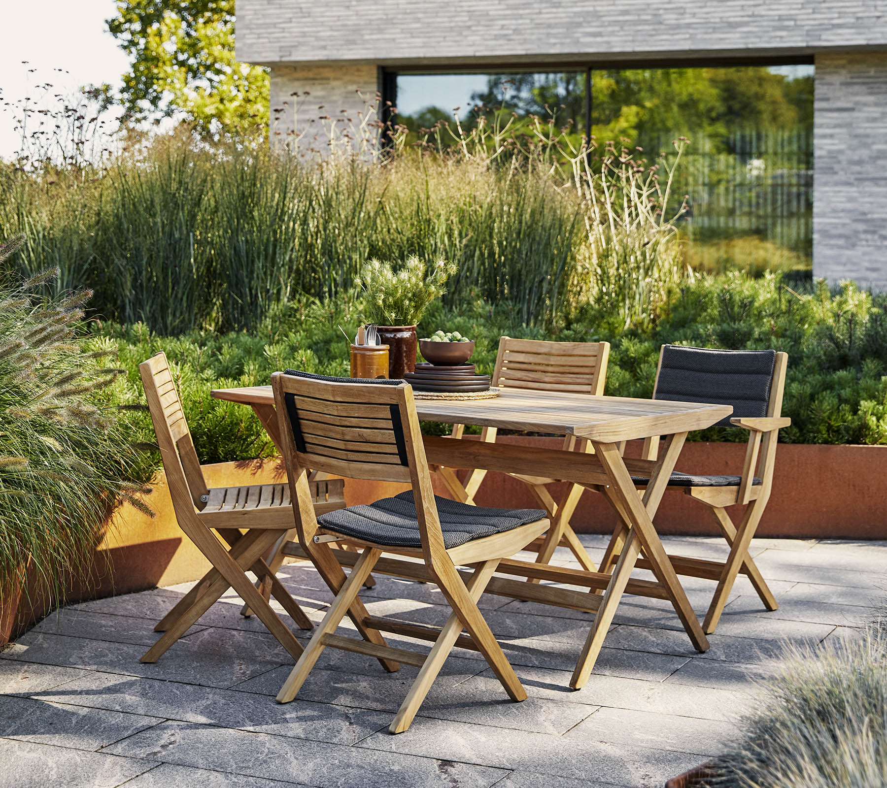 Stijlvolle houten tafel met bijpassende stoelen, omgeven door weelderig groen en een moderne architectonische achtergrond. Perfect voor buitensamenkomsten.