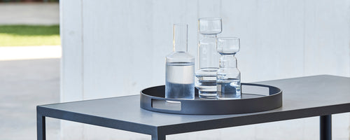 Een rond serveerblad met verschillende glazen, waaronder een hoge kan en meerdere glazen, geplaatst op een sleek tafel.