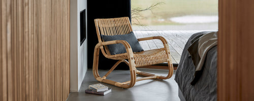 Gevlochten stoel met een comfortabele kussen, geplaatst naast een bed, dat een modern en stijlvol ontwerp in een serene omgeving toont.