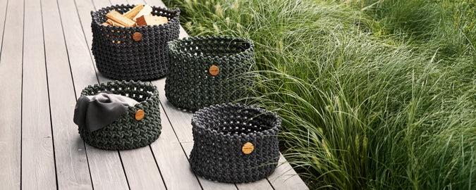 Verzameling van zwarte gevlochten manden, gerangschikt op een houten terras met groen op de achtergrond.