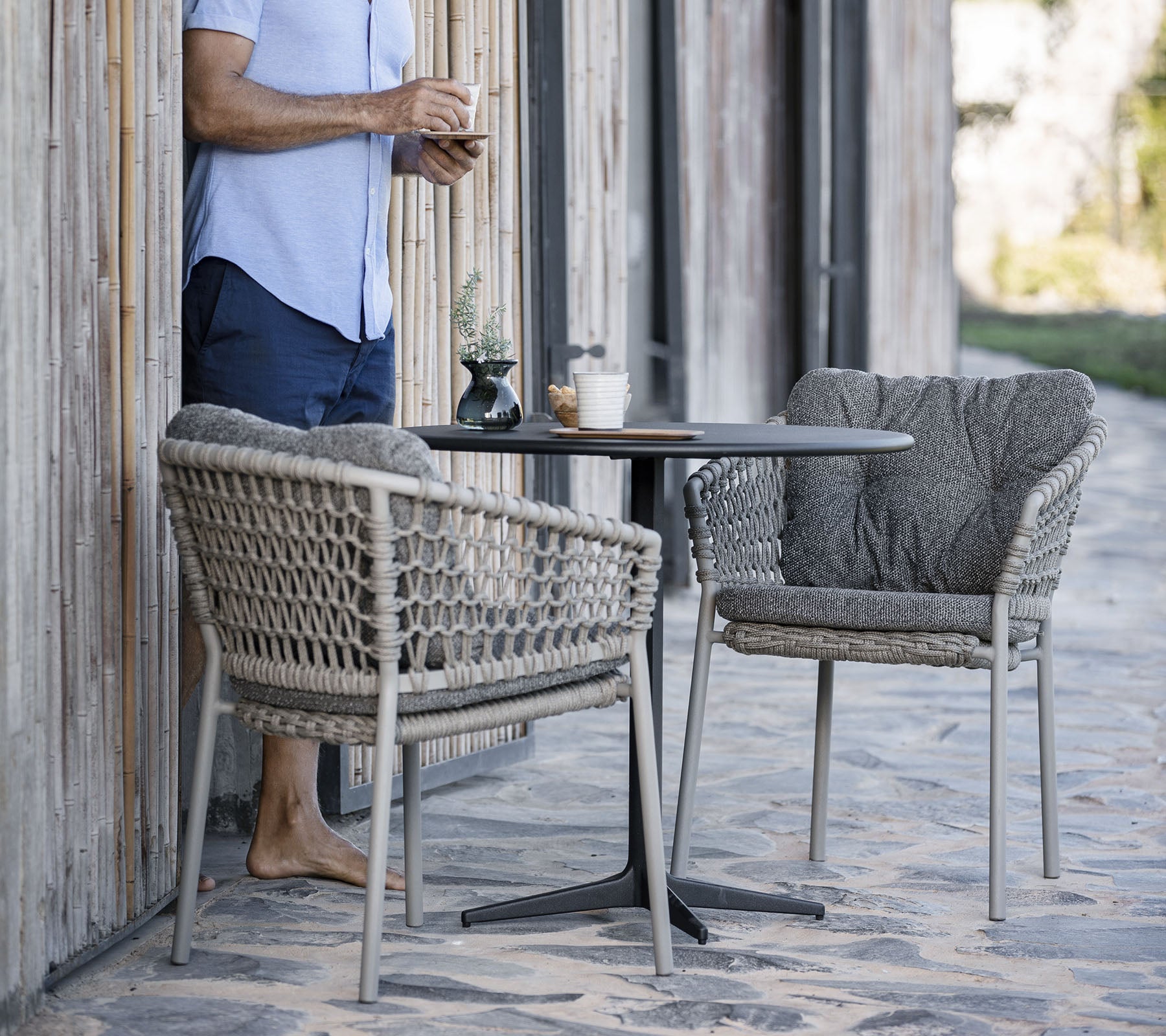 Een persoon staat naast een kleine ronde tafel met twee stijlvolle geweven stoelen en geniet van een drankje in een ontspannen omgeving.