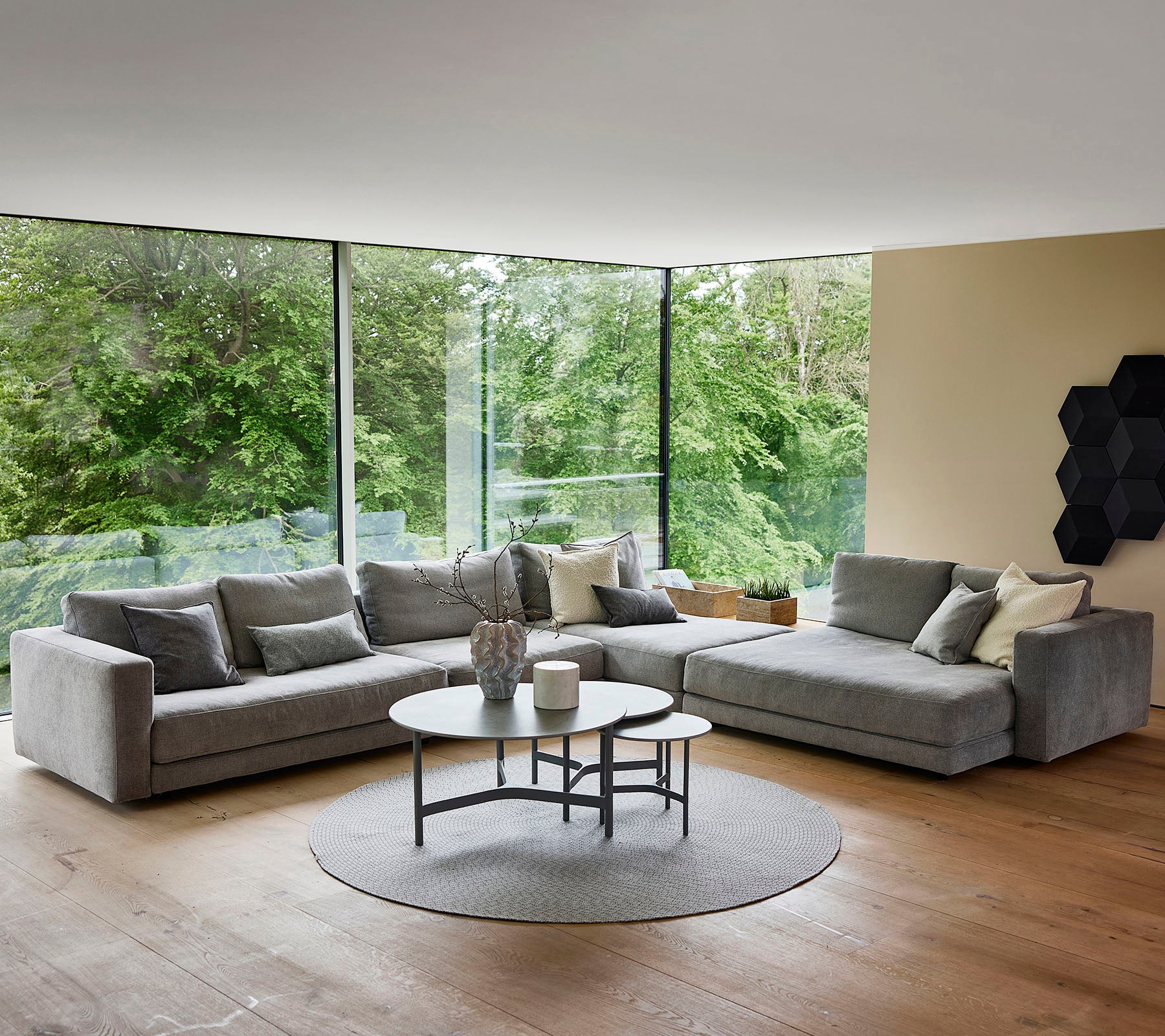 Een stijlvolle, moderne woonkamer met een pluche grijze hoekbank, een ronde salontafel en grote ramen die het groen buiten tonen.