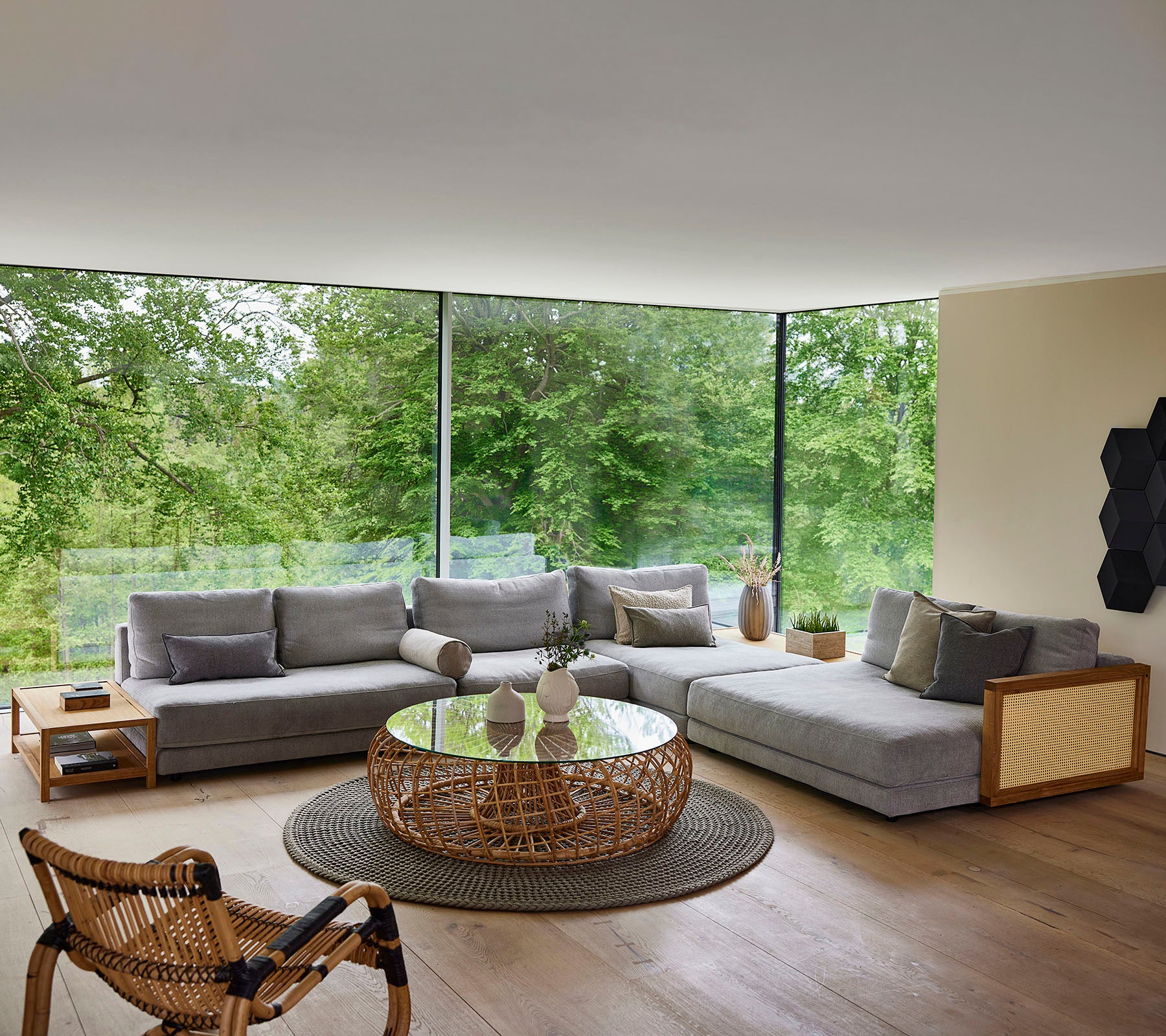 Een modern zitgedeelte met een grijze sectional bank, een ronde salontafel en een gevlochten stoel, omringd door groen.