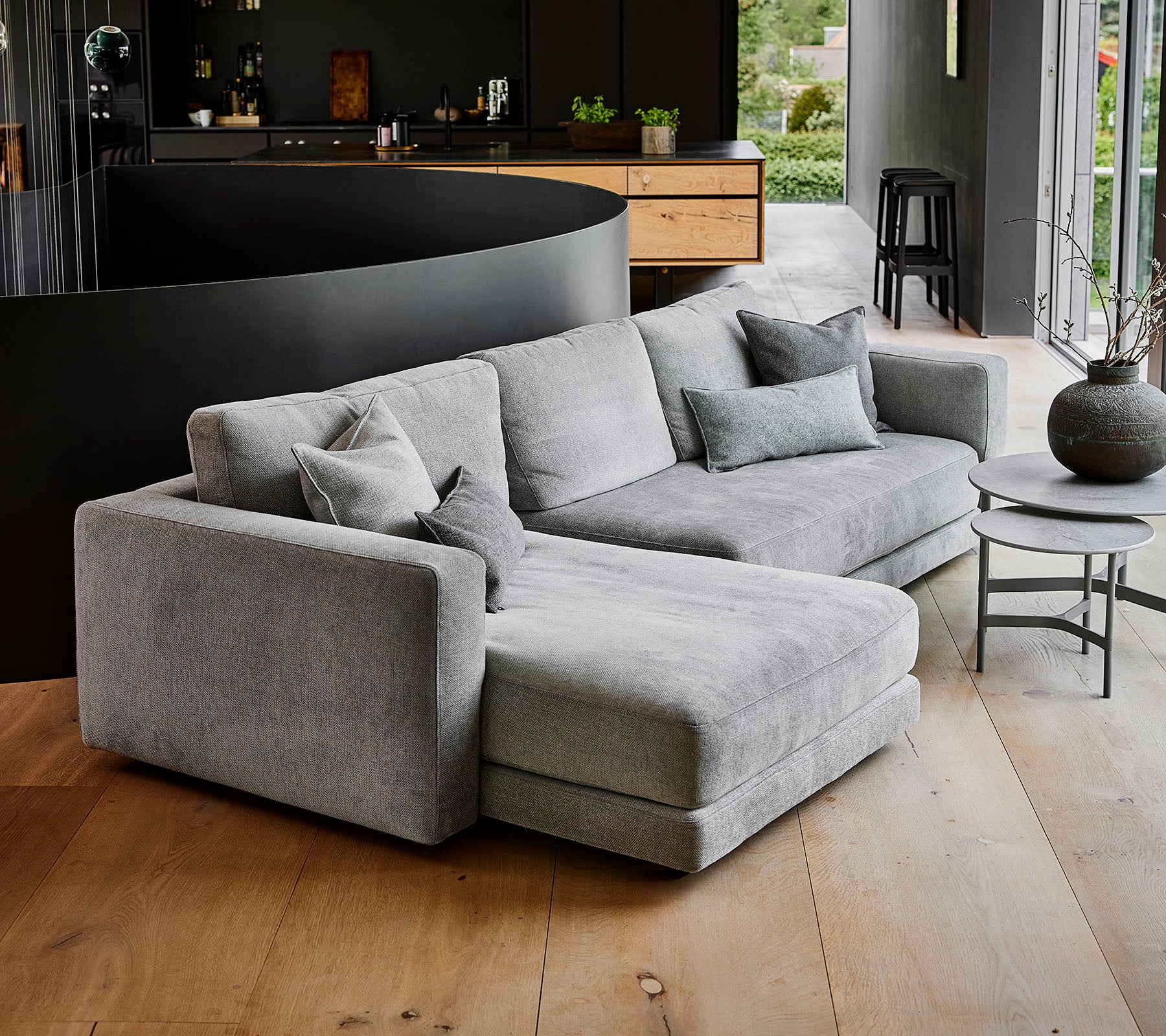 Moderne grijze hoekbank met meerdere kussens, vergezeld van een ronde salontafel en een decoratieve vaas. Eenvoudig, elegant design.