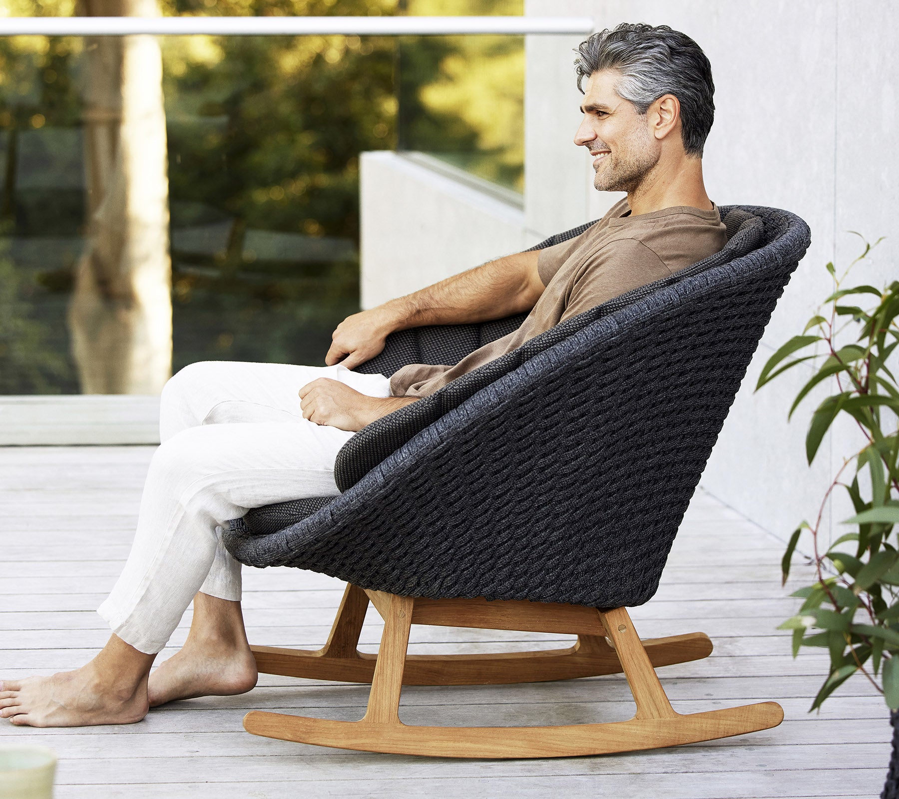 Een man zit ontspannen in een stijlvolle, moderne schommelstoel met een geweven design en een basis van natuurlijk hout, omringd door groen.