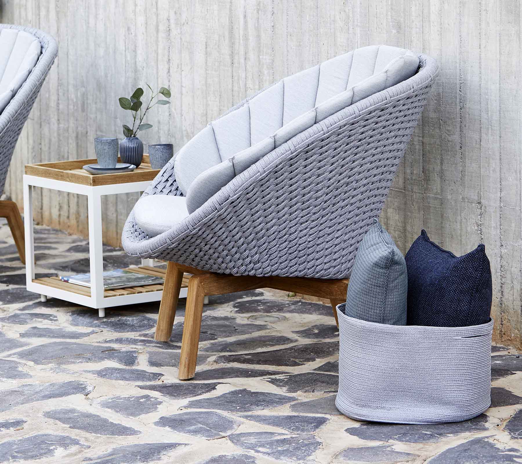 Moderne grijze loungestoel met een geweven ontwerp, vergezeld van kussens en een klein houten bijzettafeltje. De stenen vloer verbetert de esthetiek.