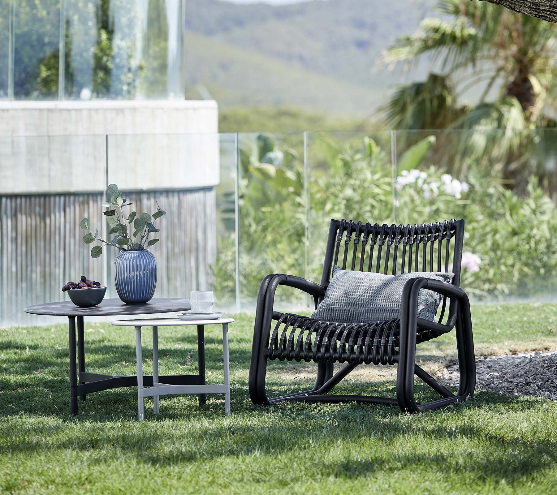 Moderne loungechair met een gemêleerd ontwerp, vergezeld van een bijzettafel met een vaas en een kamerplant, geplaatst in een natuurlijke omgeving.