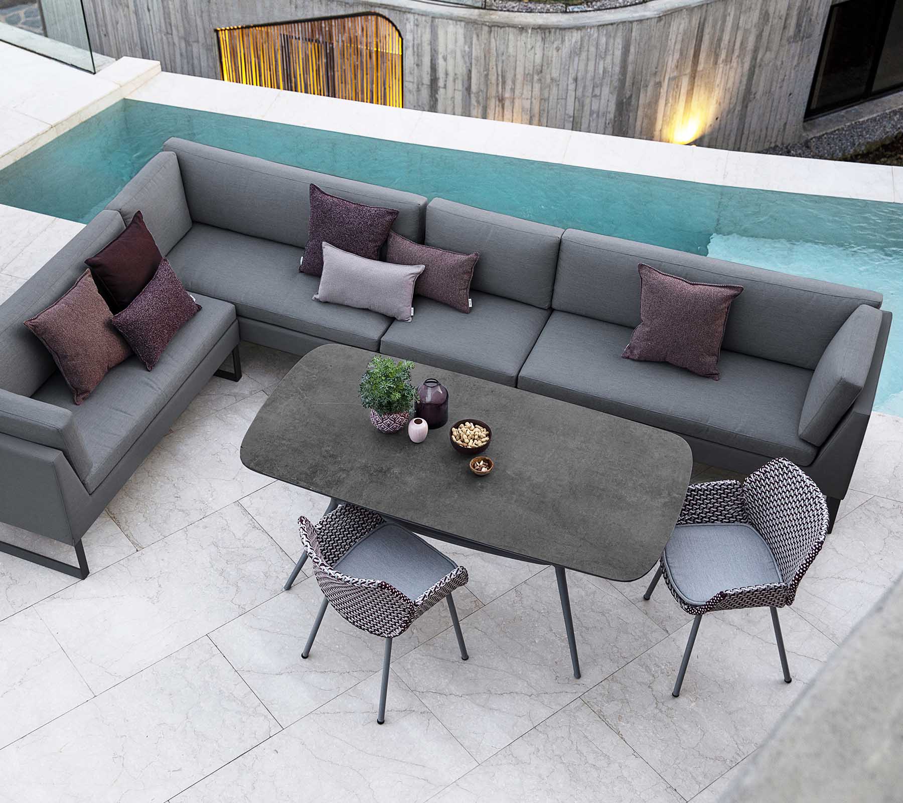 Moderne buitenzitarrangement met een grijze hoekbank, decoratieve kussens en een strak eettafel omringd door stijlvolle stoelen.