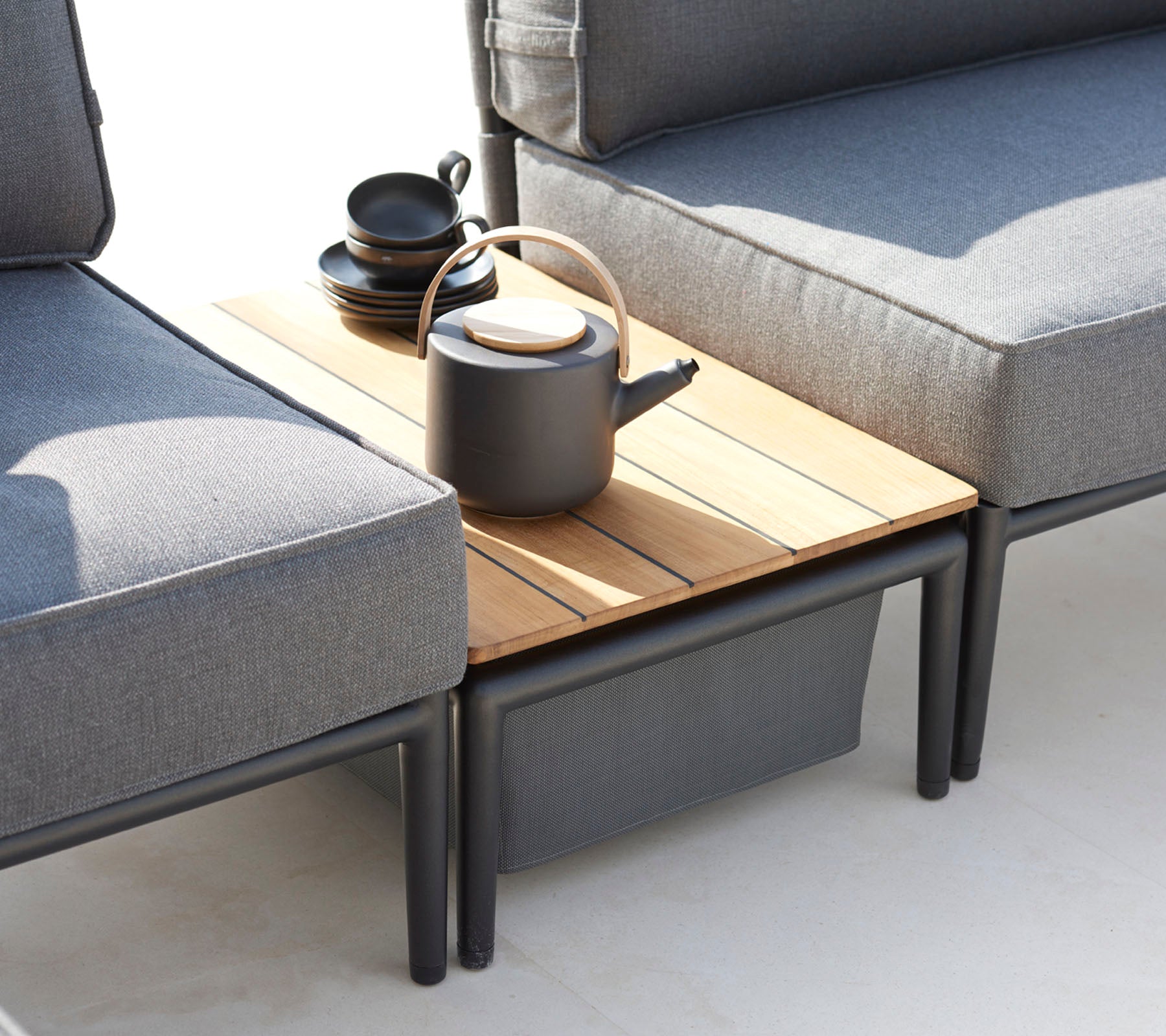 Twee grijze banken flankeren een houten bijzettafel, bekleed met een zwarte theepot en verschillende kopjes, wat een gezellige zithoek creëert.