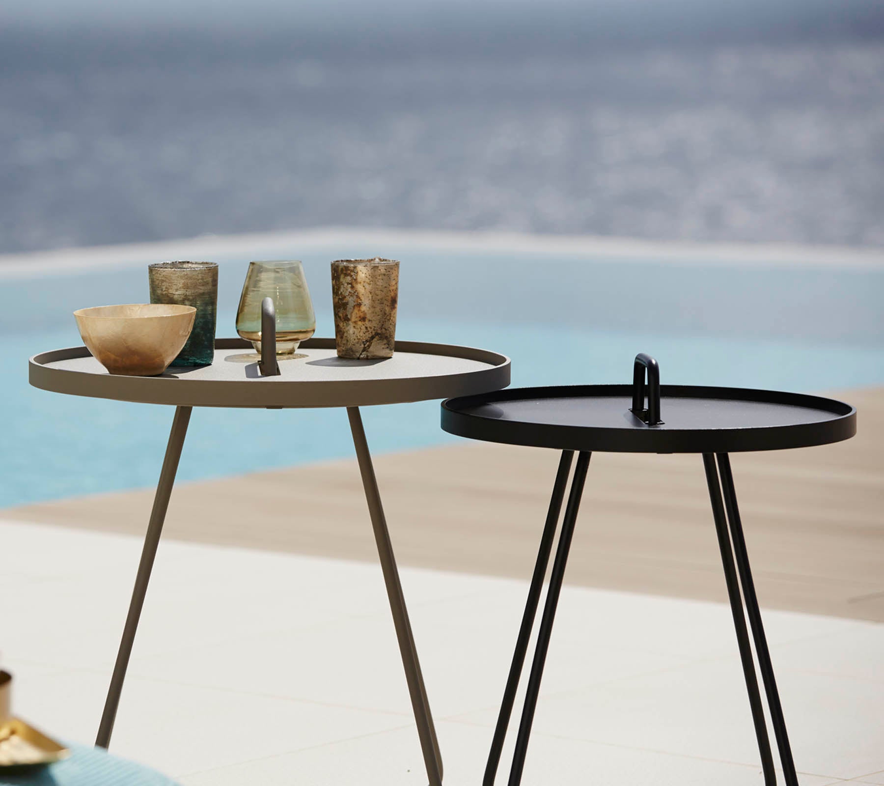 Twee stijlvolle bijzettafels met verschillende decoratieve glazen op de top, geplaatst bij een serene waterelement op de achtergrond.