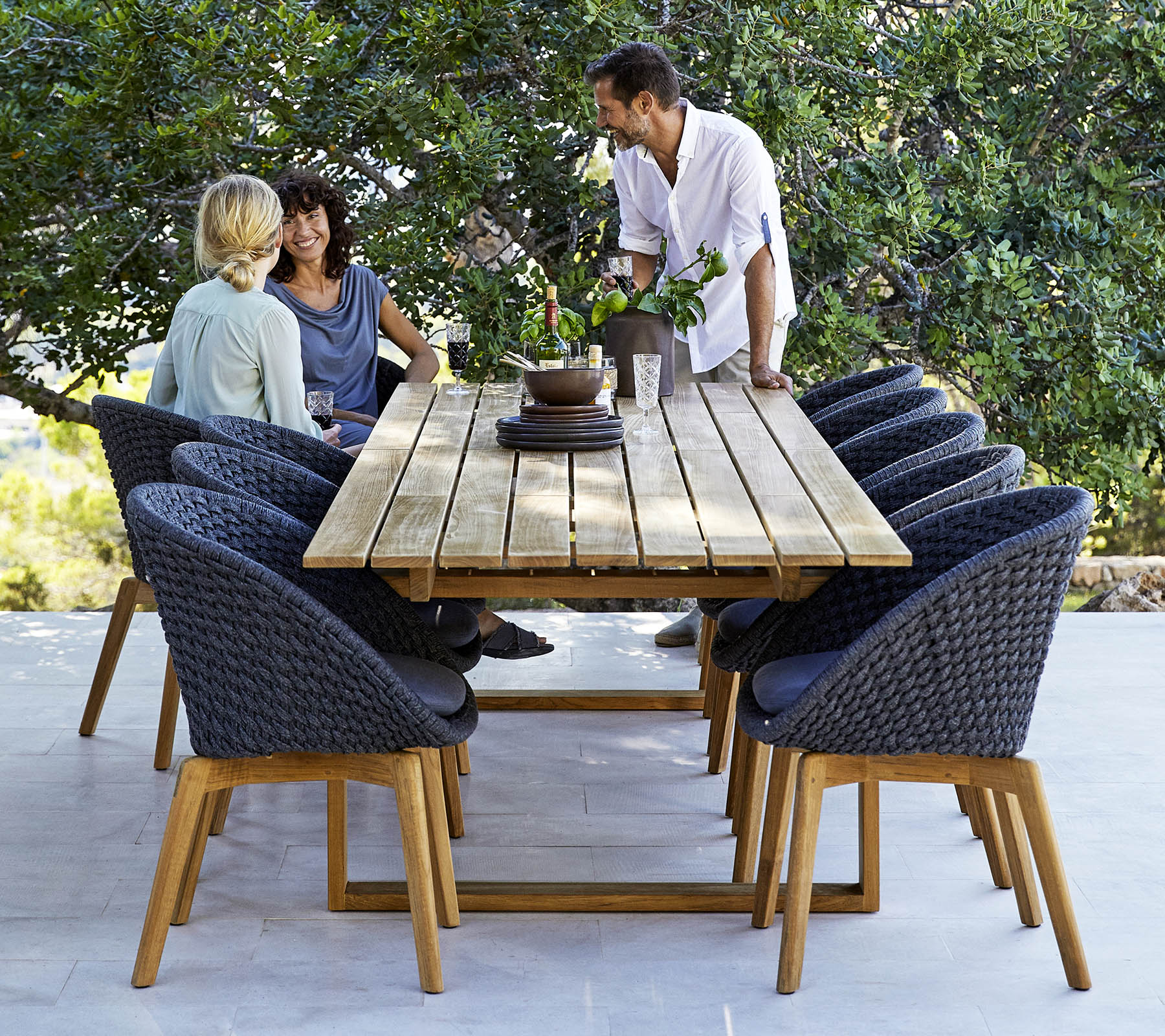 Een stijlvolle eetopstelling met een houten tafel omringd door comfortabele stoelen, met drie mensen die zich in een gesprek verdiepen, te midden van groen.