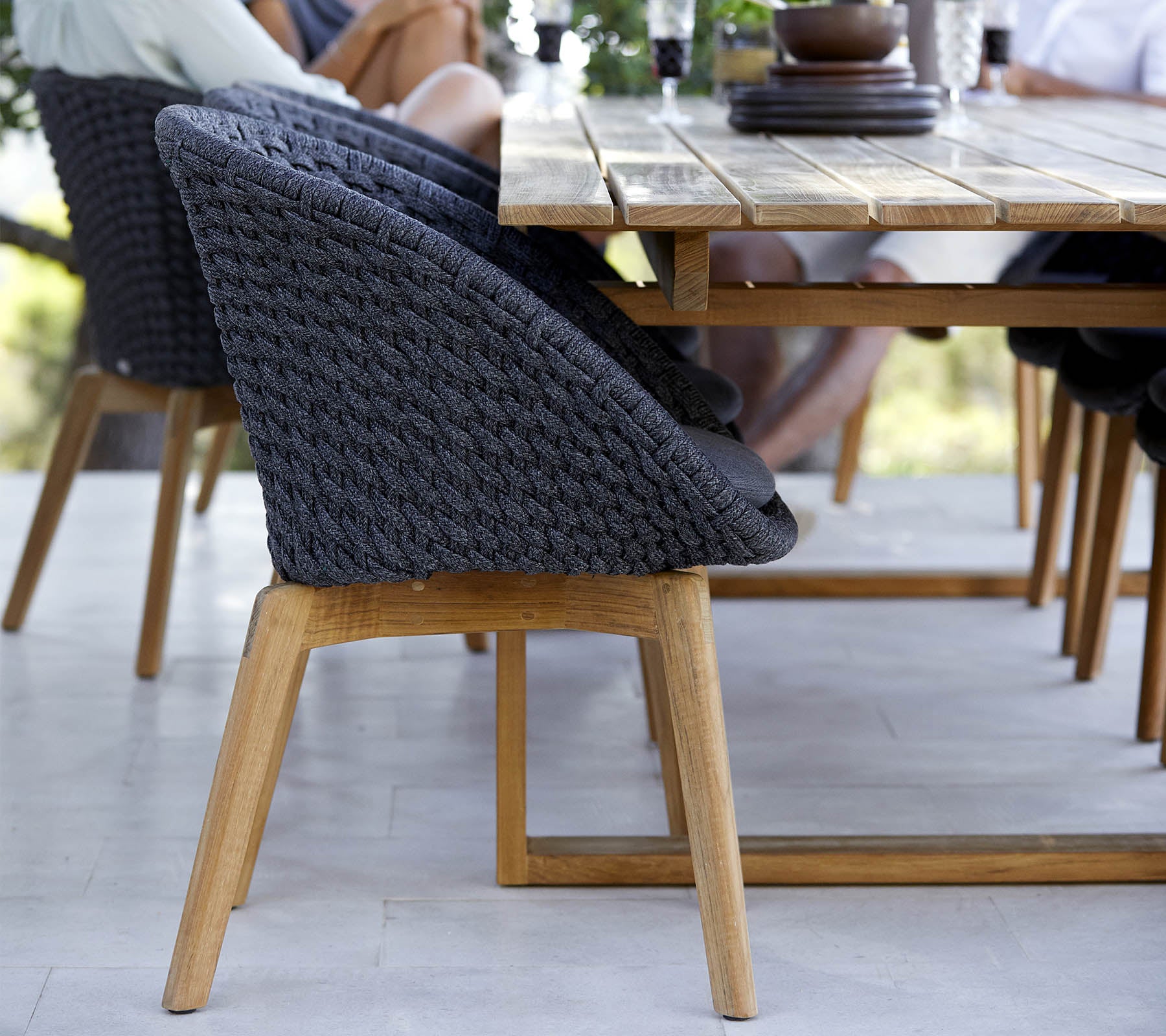 Een stijlvolle stoel met een gevlochten rugleuning en houten poten, naast een houten eettafel, met mensen in de buurt die bezig zijn.