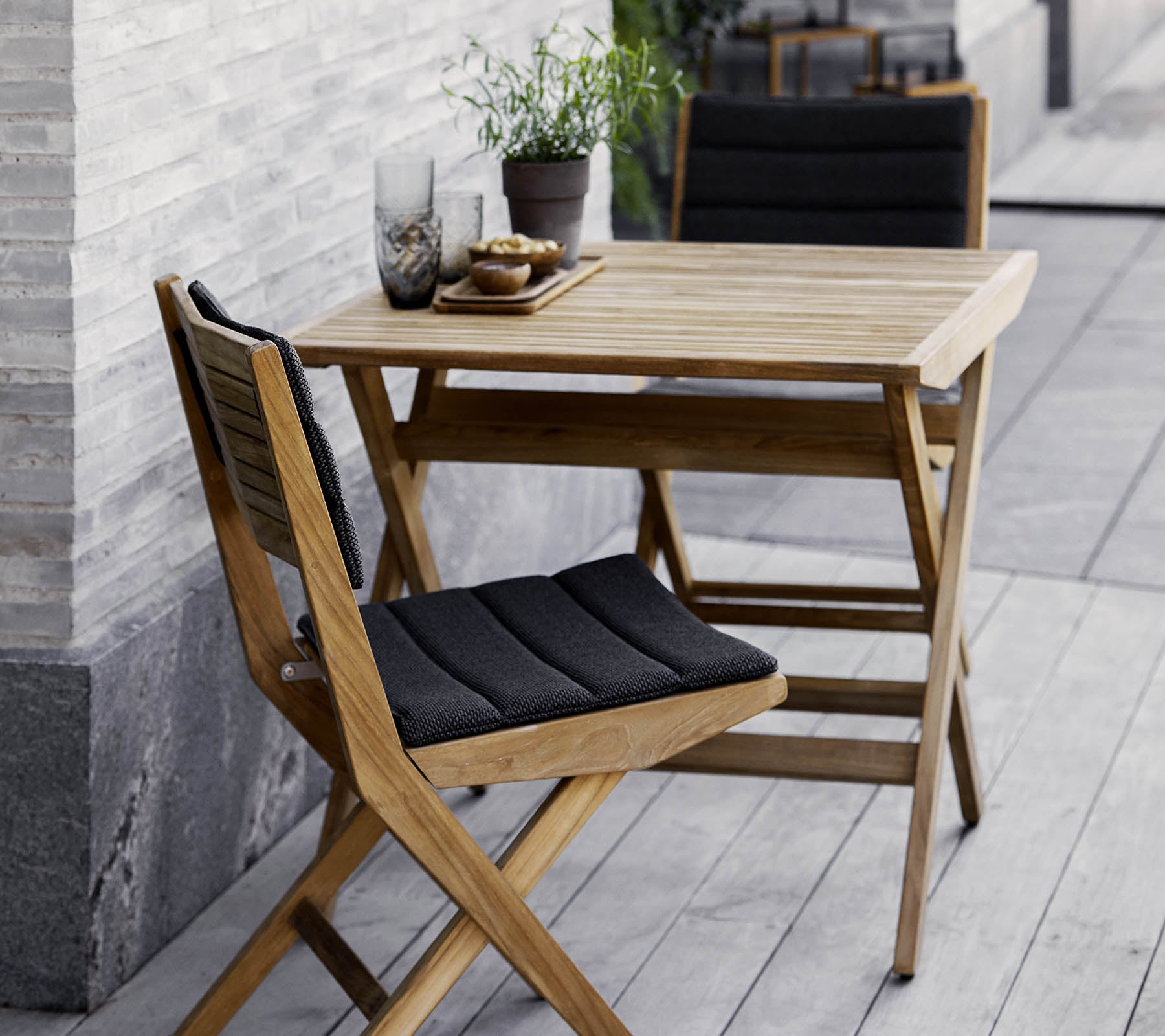Houten tafel en stoelen set met zwarte kussens, versierd met potplanten en decoratief glaswerk. Perfect voor een gezellige setting.