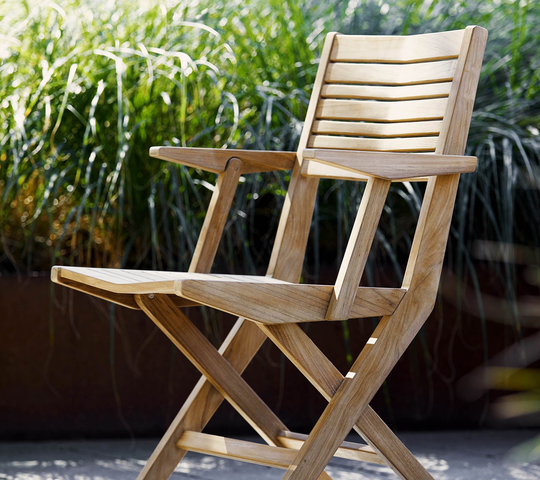 Een houten opvouwbare stoel met latten en armleuningen, omringd door weelderig groen loof. Ideaal voor ontspanning en buitenomgevingen.