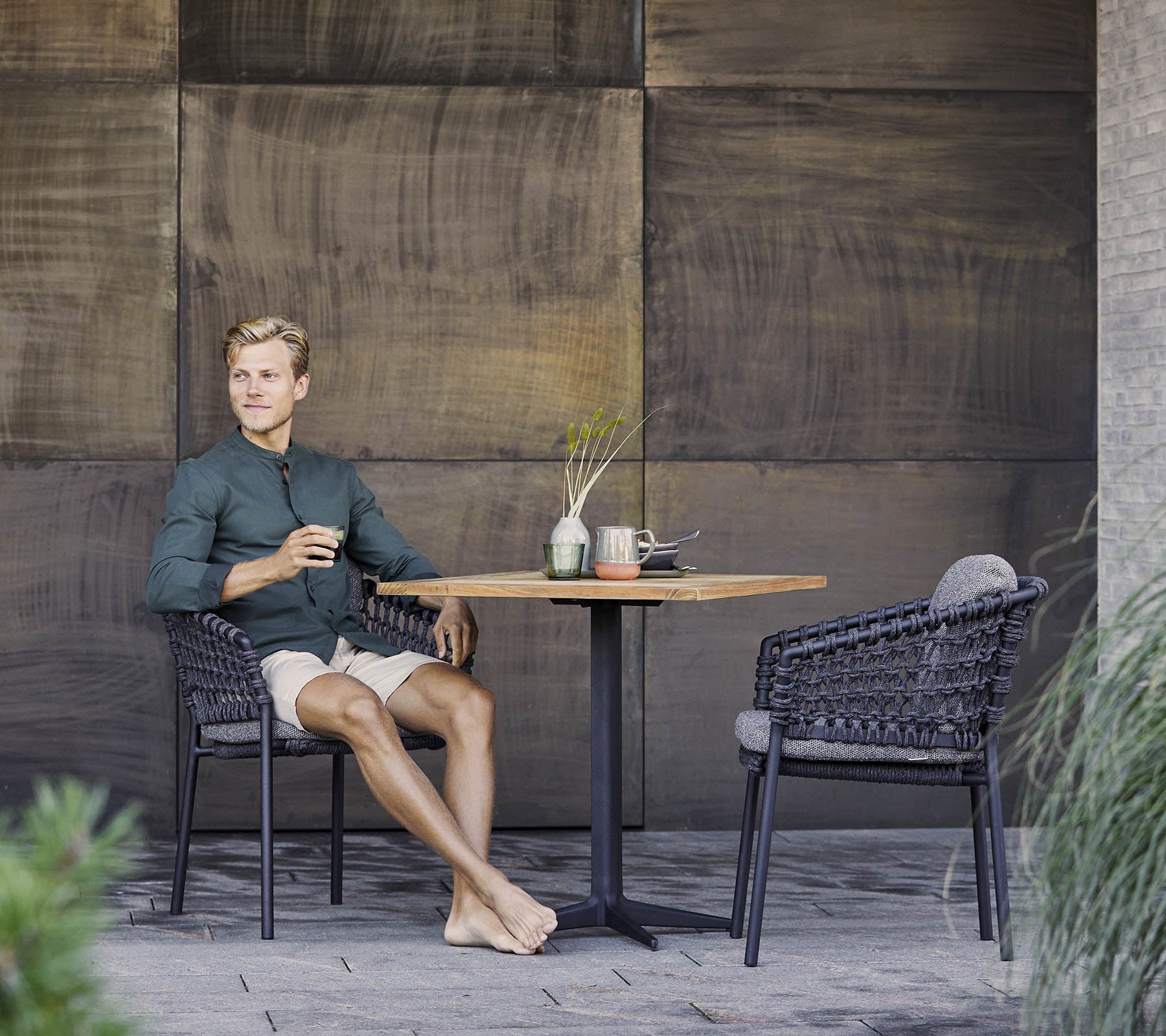 Een persoon zit ontspannen aan een tafel, geniet van een drankje, omringd door stijlvolle rotan stoelen en decoratieve elementen.