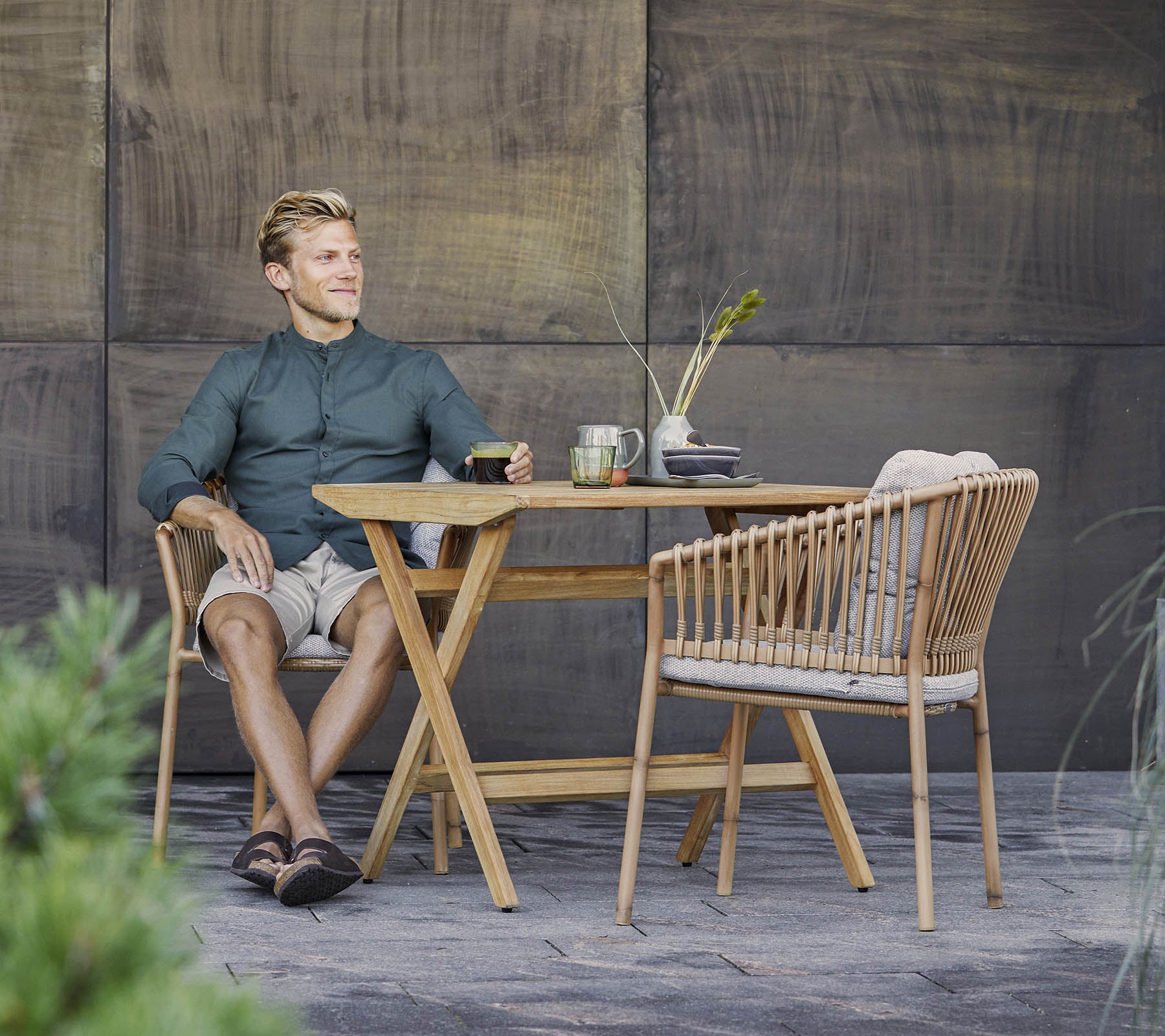 Een man zit ontspannen aan een stijlvolle houten tafel met een lichte drank, omringd door groen en een gestructureerde achtergrond.