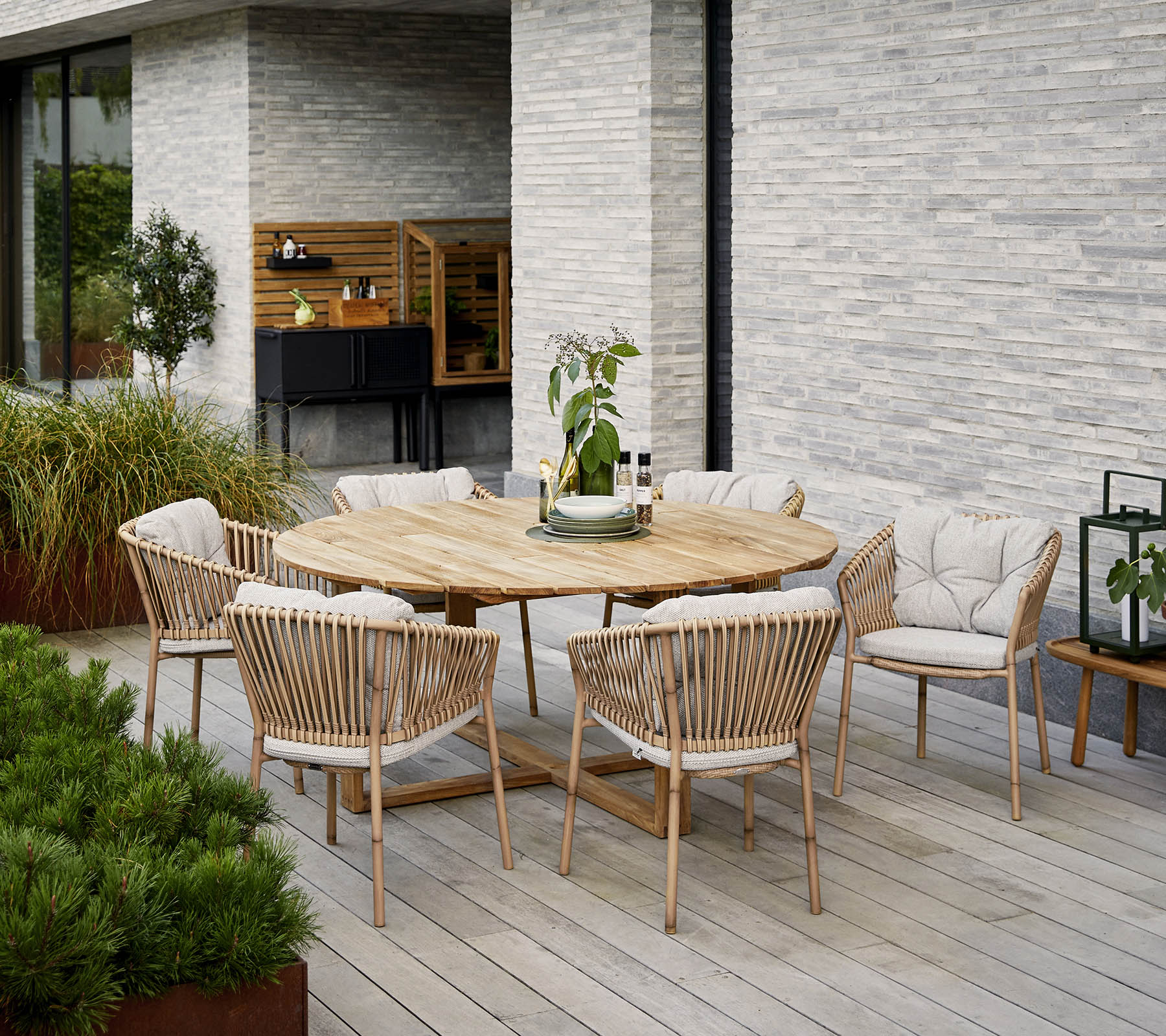 Ronde houten eettafel omringd door gestoffeerde stoelen, met een decoratief plantenmiddelstuk, tegen een gestructureerde stenen muur.