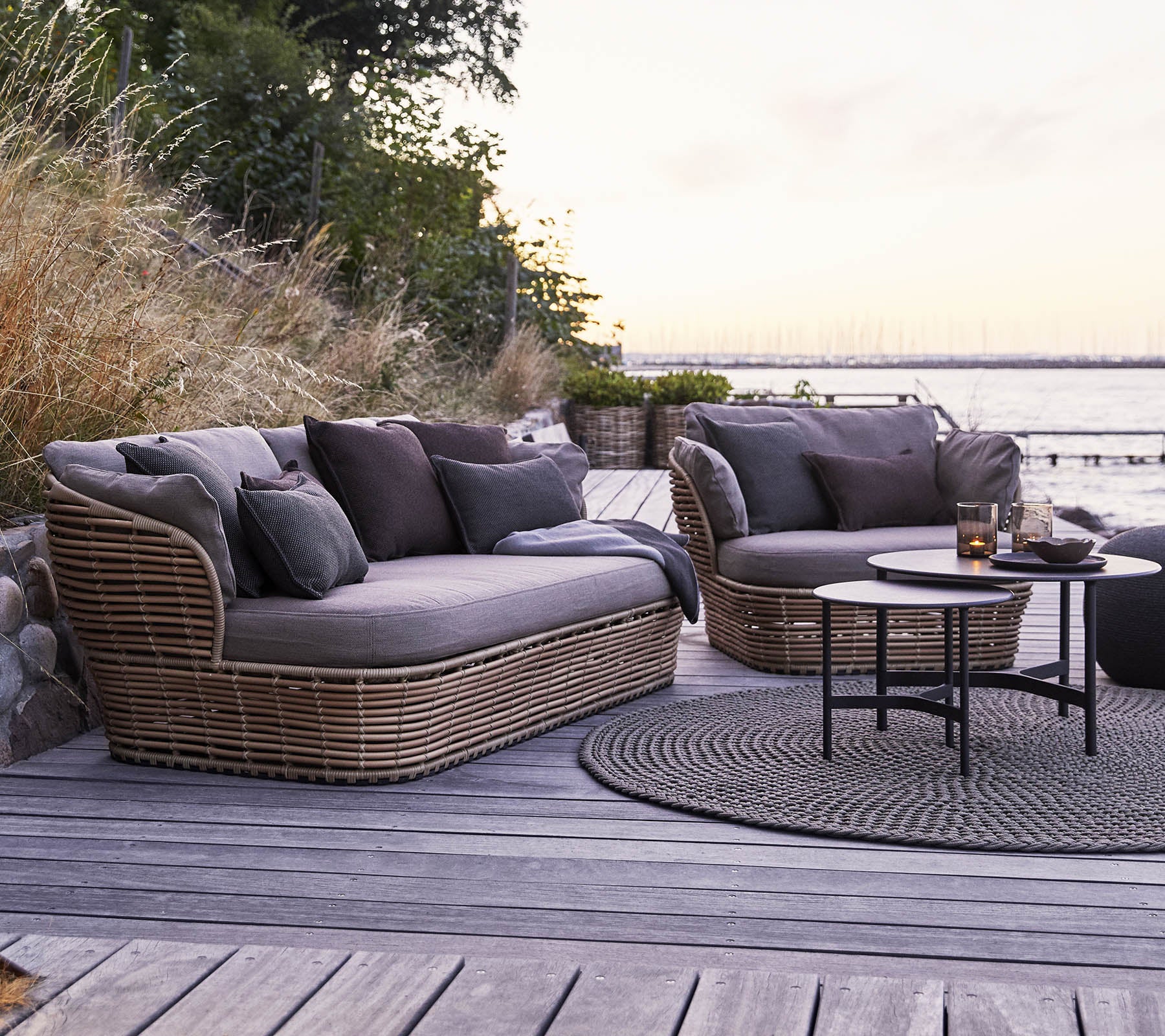 Stijlvolle buitenseating met twee zachte, gebogen sofas met donkere kussens en een ronde salontafel op een houten oppervlak.