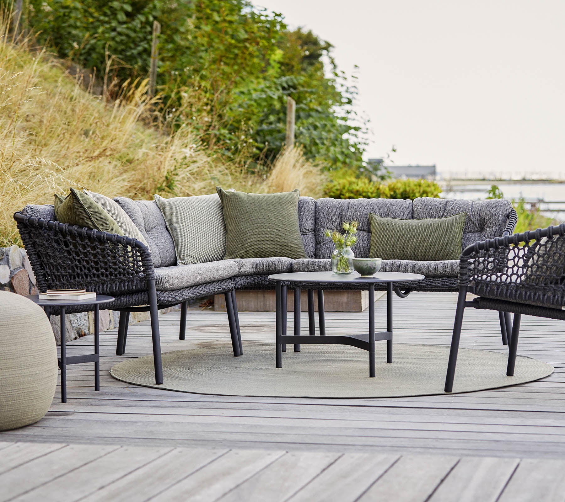 Stijlvolle buitenzitarrangement met een gebogen hoekbank, ronde tafel en gezellige accenten in een natuurlijke omgeving.