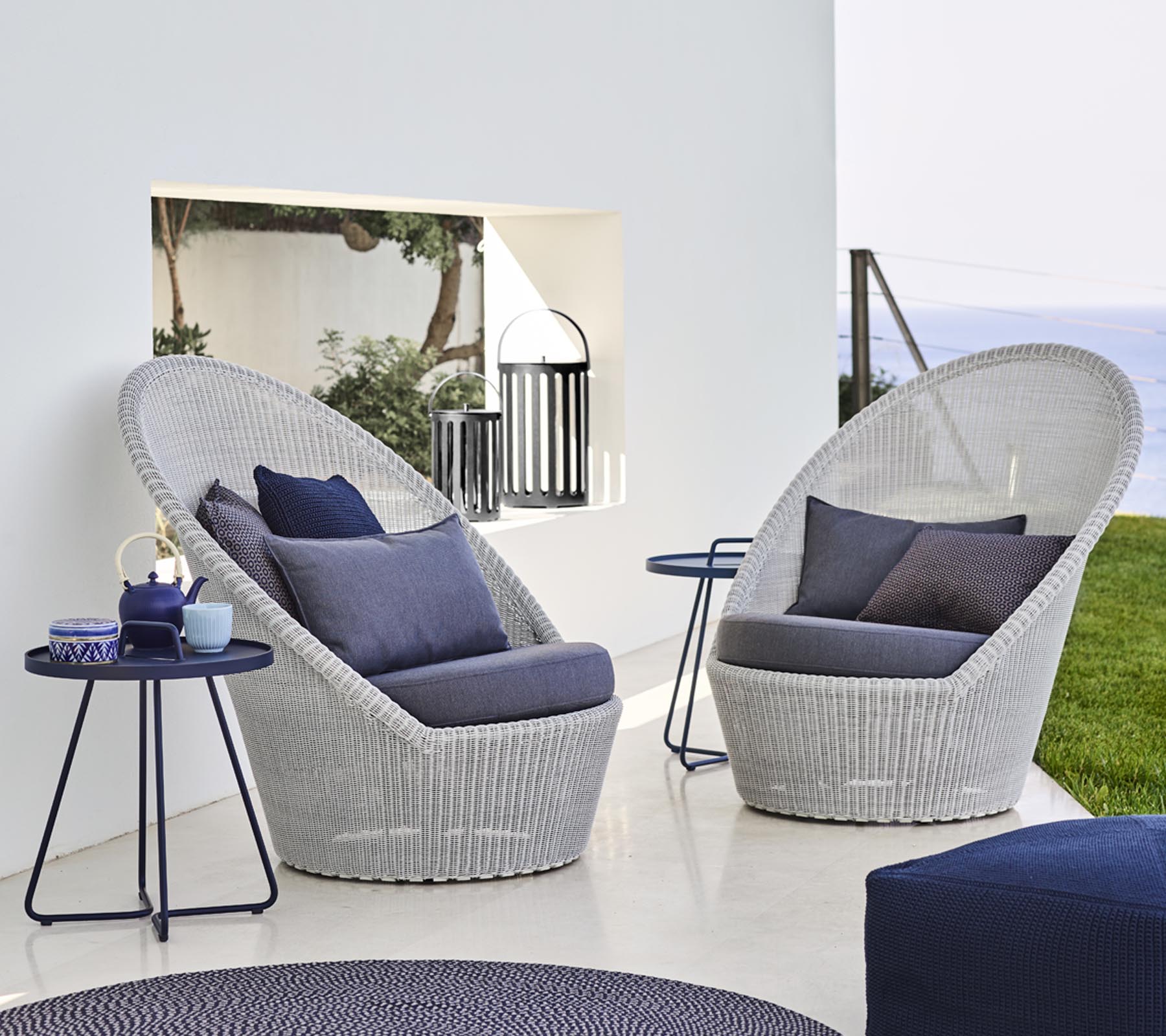 Twee stijlvolle gevlochten loungestoelen met blauwe kussens naast een tafeltje, tegen een serene achtergrond.