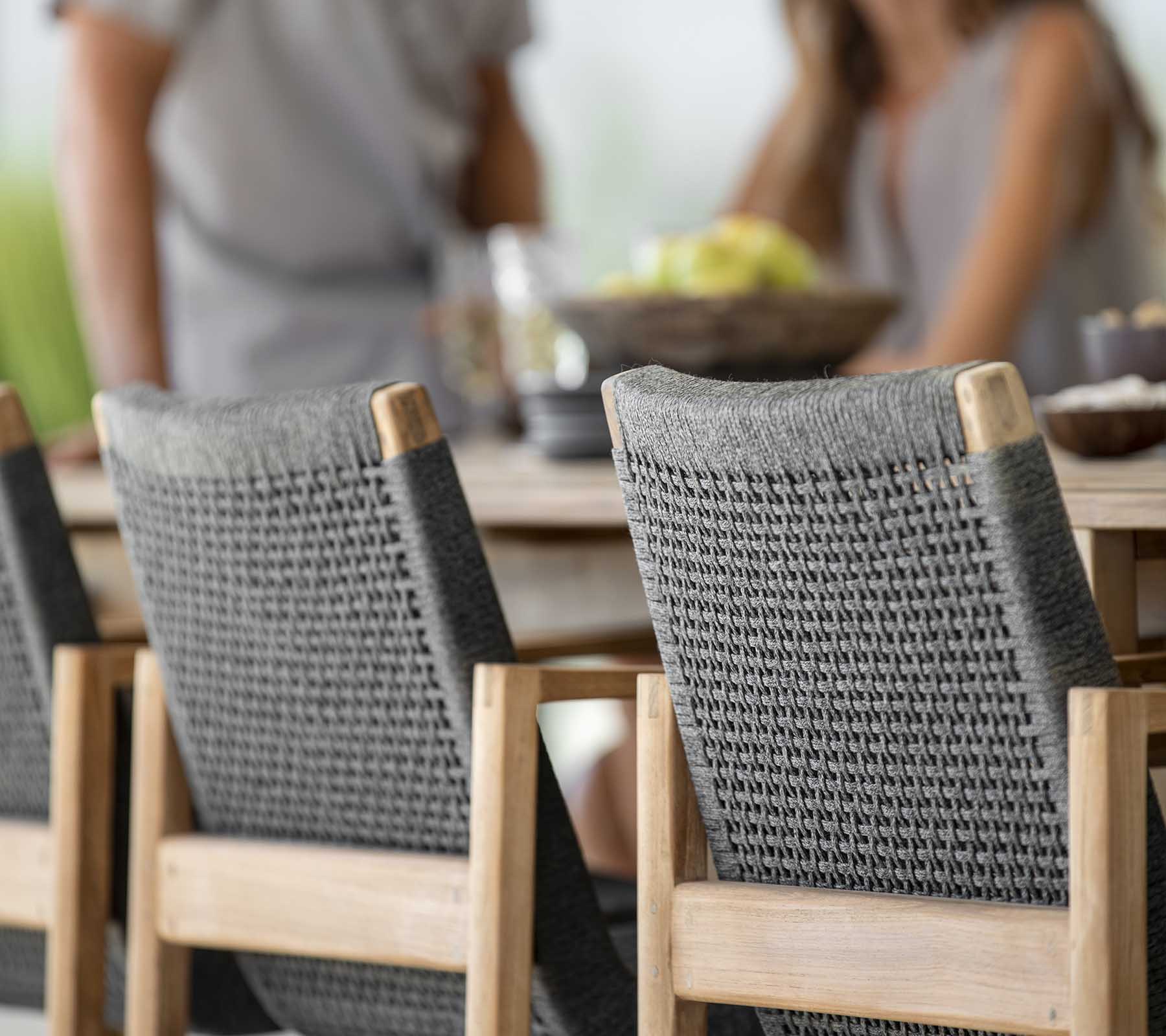 Buiteneet scène met een houten tafel en twee stijlvolle grijze gevlochten stoelen, waar mensen samen van een maaltijd genieten.