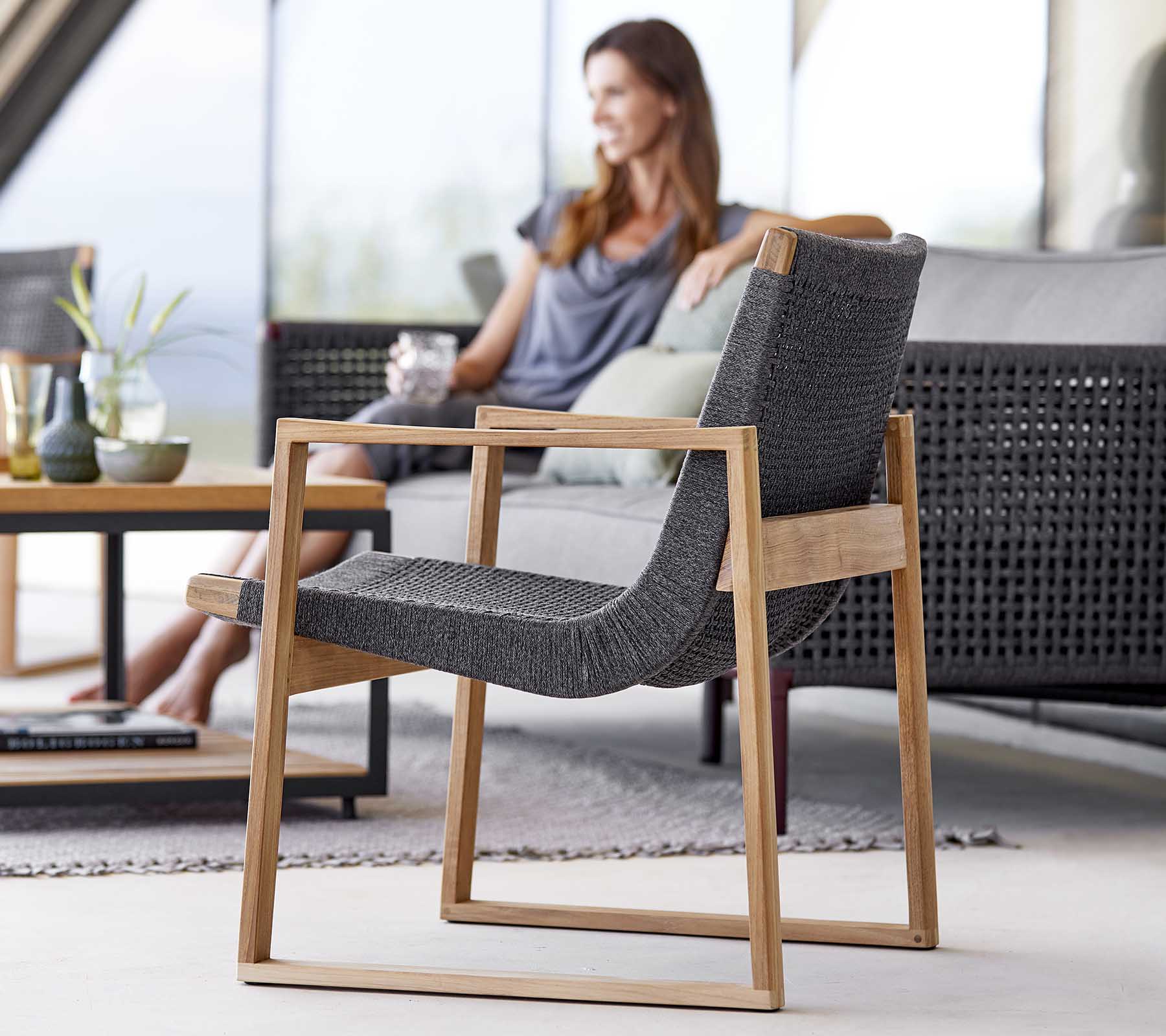 Een moderne stoel met een slanke houten frame en geweven stof, naast een vrouw die ontspant en geniet van een drankje.