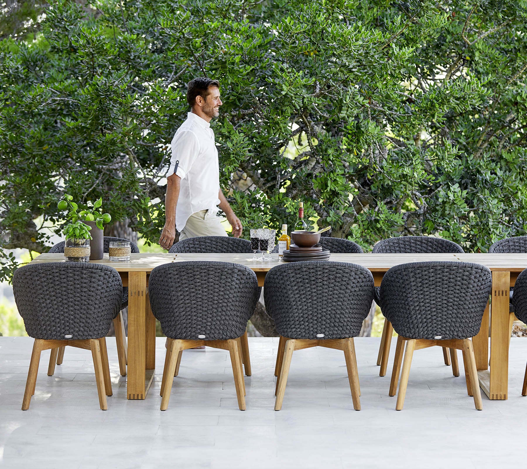 Een man loopt voorbij een grote houten eettafel omringd door moderne, gevlochten stoelen en groen op de achtergrond.