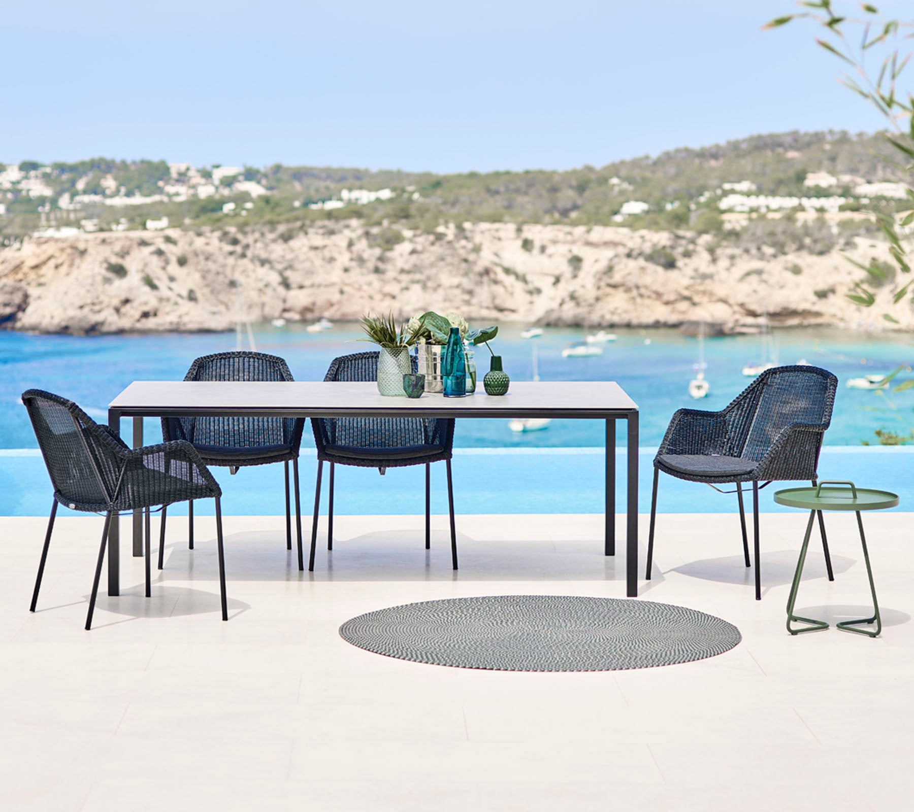 Een moderne buiten eettafelset met een rechthoekige tafel, geweven stoelen en een klein bijzettafeltje, met een schilderachtige strandachtergrond.