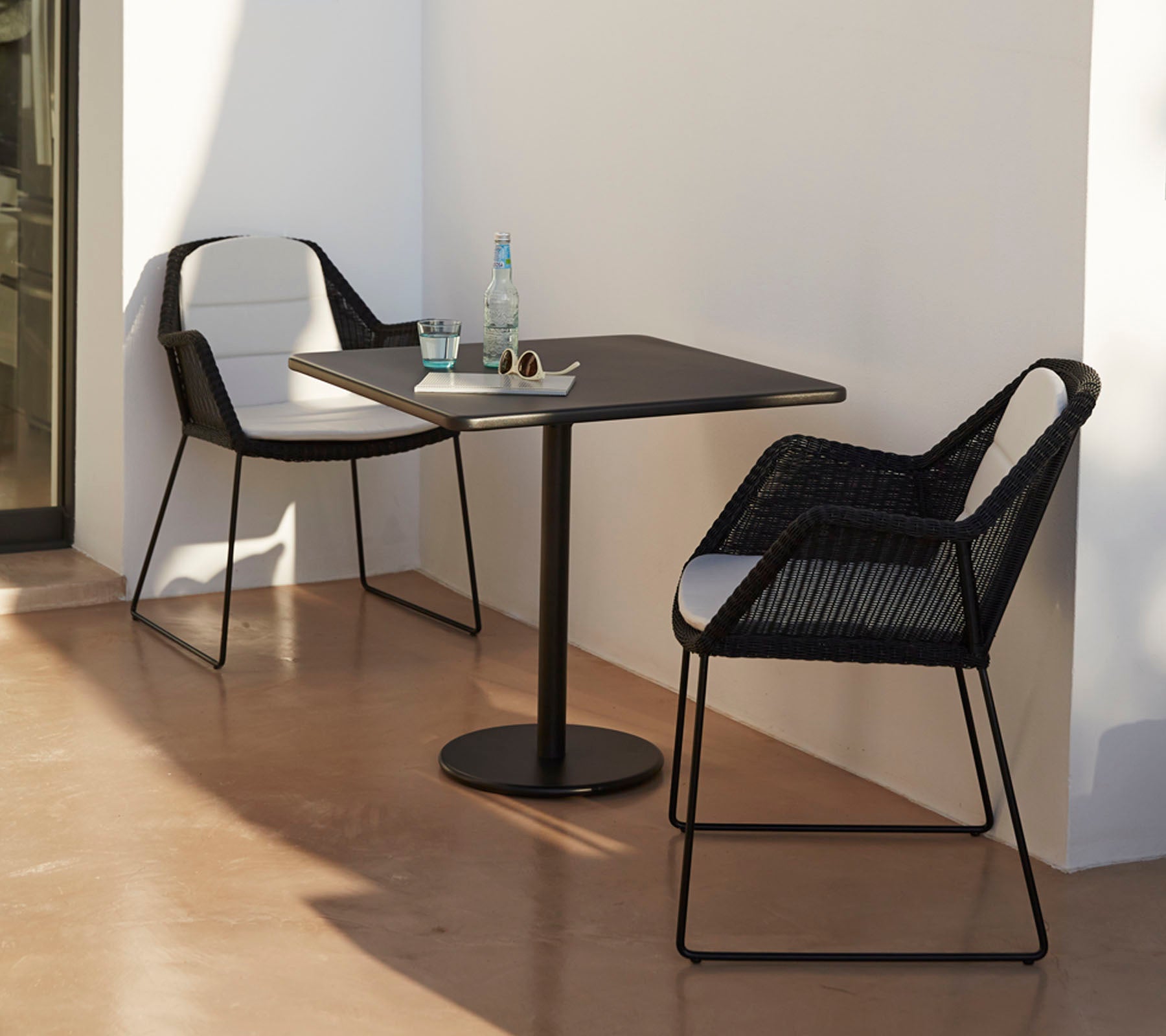 Modern tafel met twee stijlvolle zwarte stoelen, vergezeld van een waterfles en zonnebril, tegen een minimalistische achtergrond.