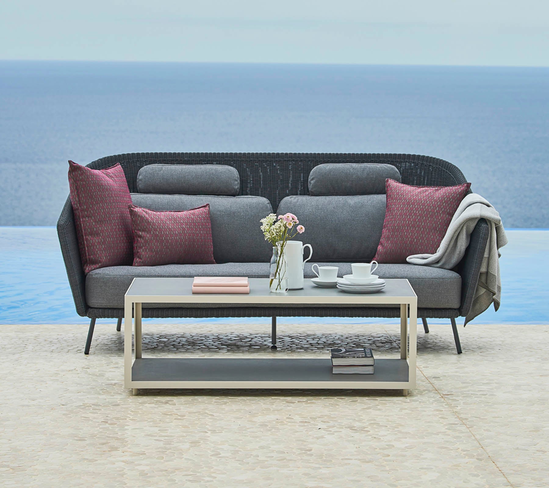 Stijlvolle grijze sofa met decoratieve kussens en een gezellige plaid, aangevuld met een strak salontafeltje met een theepot en boeken, tegen een serene achtergrond.