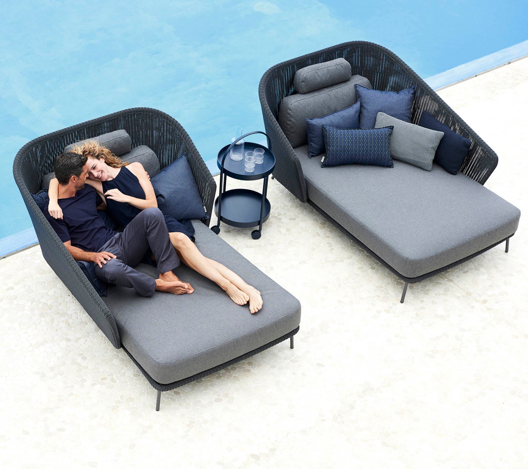 Een stel ontspant samen op stijlvolle ligstoelen met pluche kussens, vergezeld van een klein bijzettafel en decoratieve kussens.