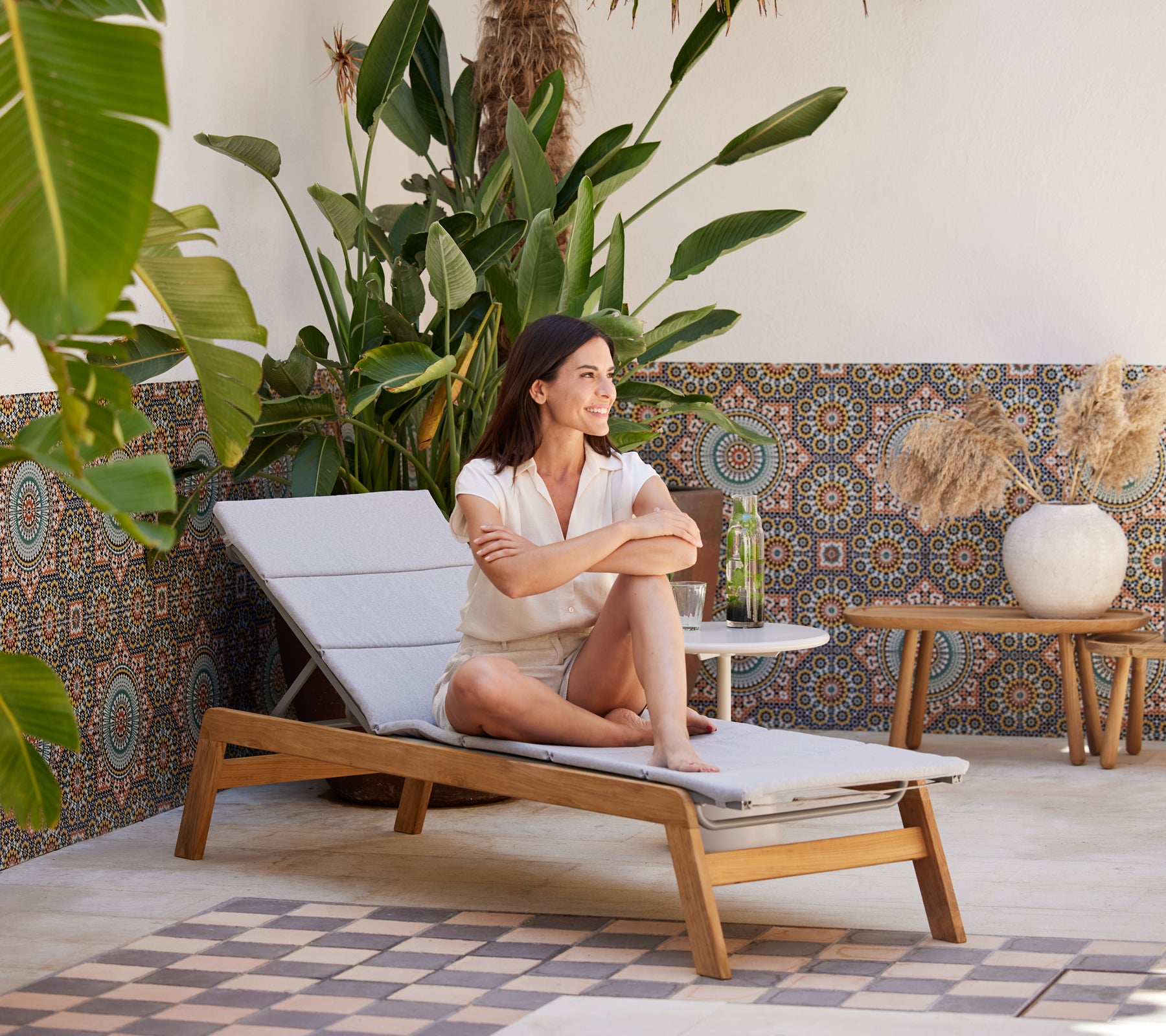 Een vrouw ontspant op een chaise longue omringd door levendige planten en met patronen versierde decoraties, terwijl ze geniet van een verfrissend drankje.
