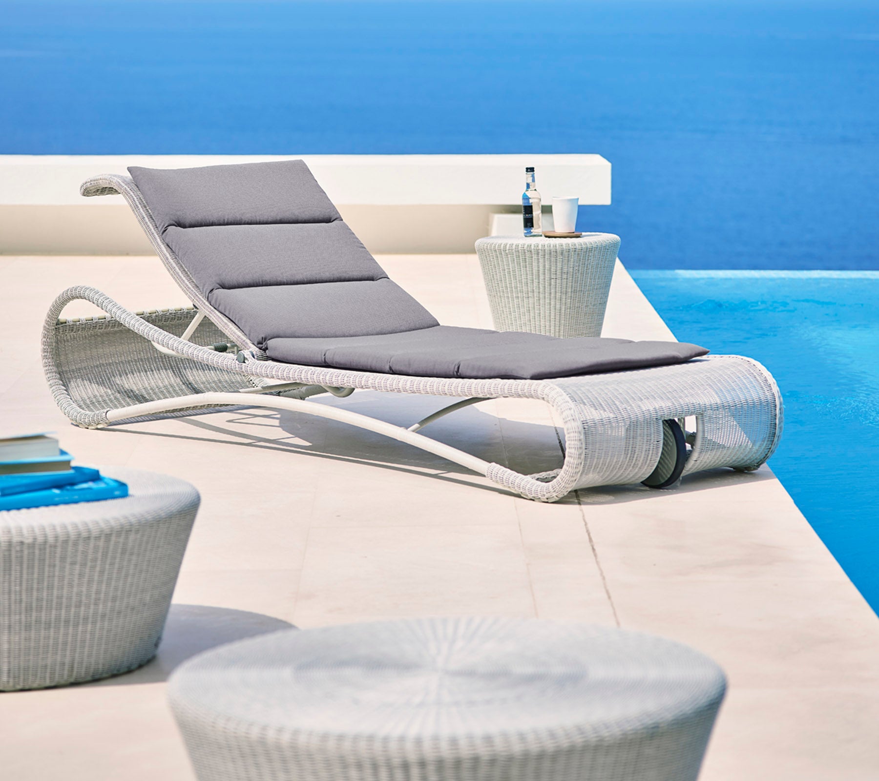 Stijlvolle loungechair met grijze kussen naast moderne sidetafels, tegen een serene blauwe achtergrond. Perfect voor ontspanning.
