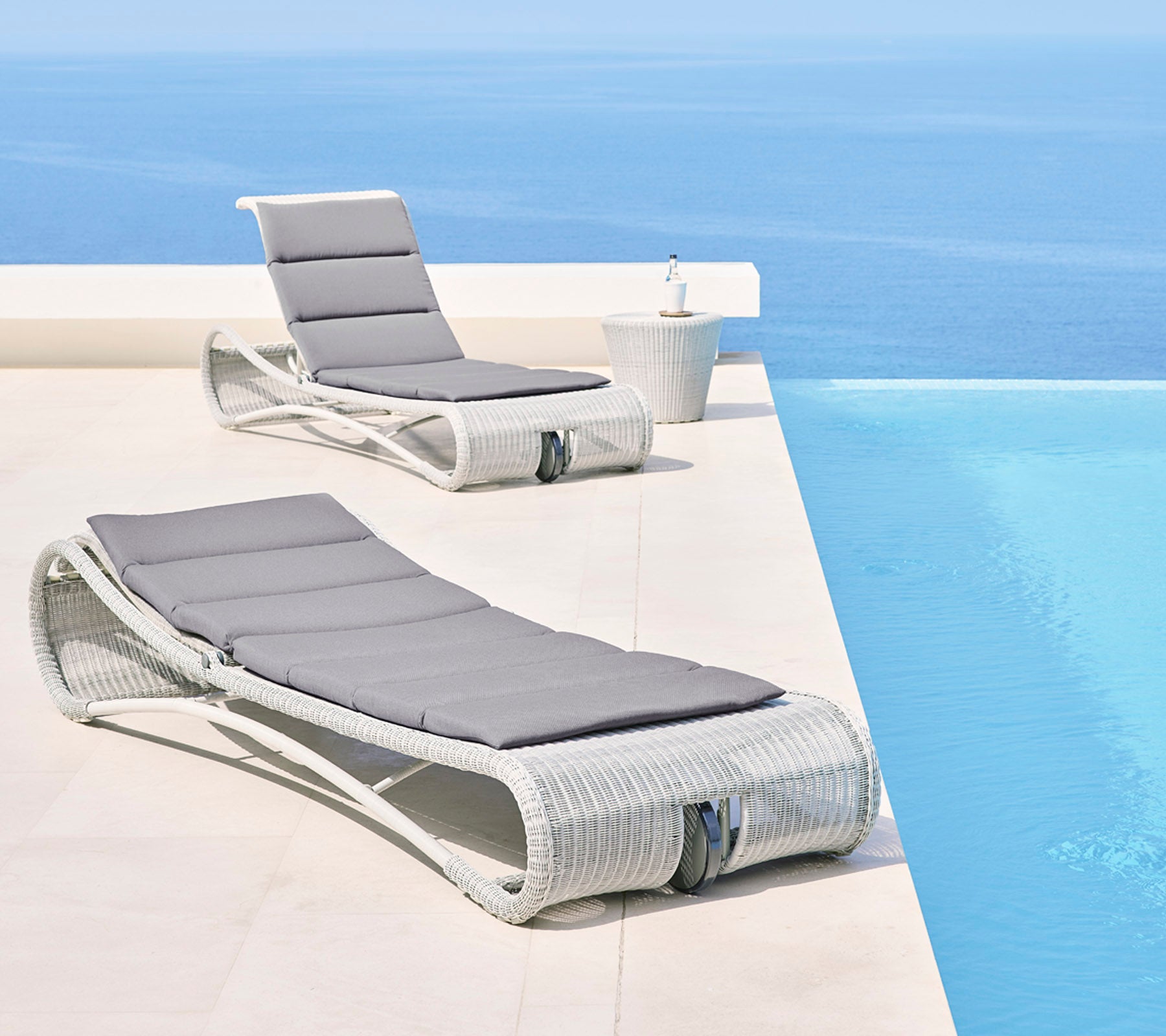 Twee stijlvolle loungefauteuils met grijze kussens naast een serene blauwe infinitypool, wat een ontspannende buitenruimte creëert.