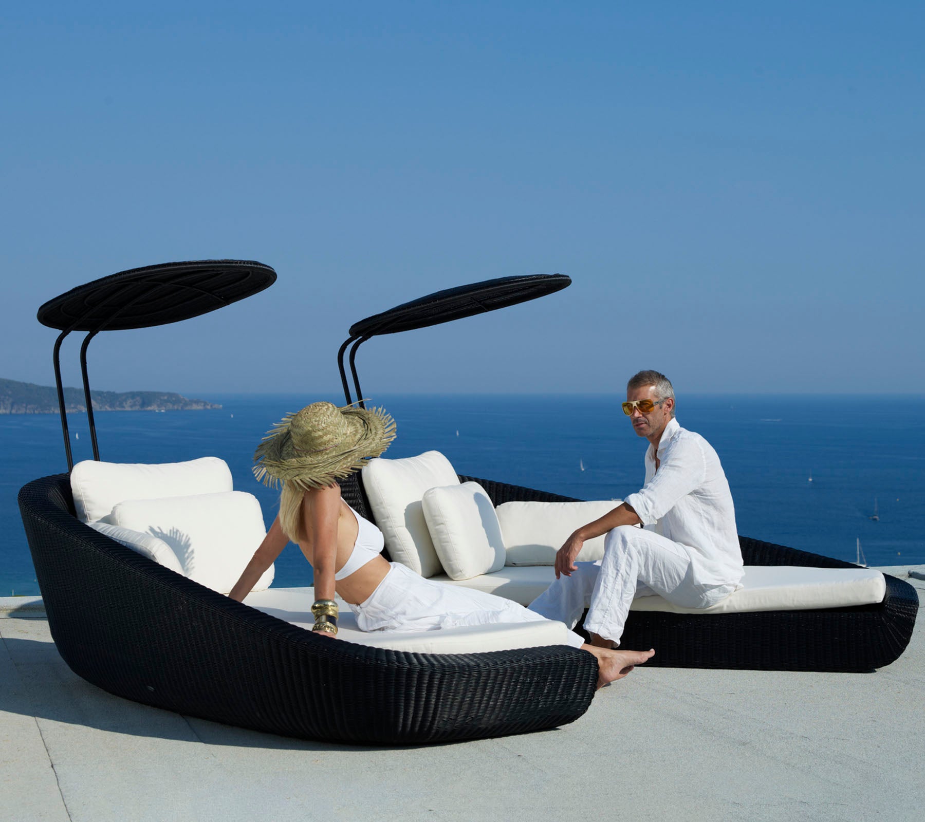 Twee mensen ontspannen op een stijlvolle, gebogen zonnebed met een schilderachtig uitzicht op de oceaan, met een strak zwart design en zachte witte kussens.